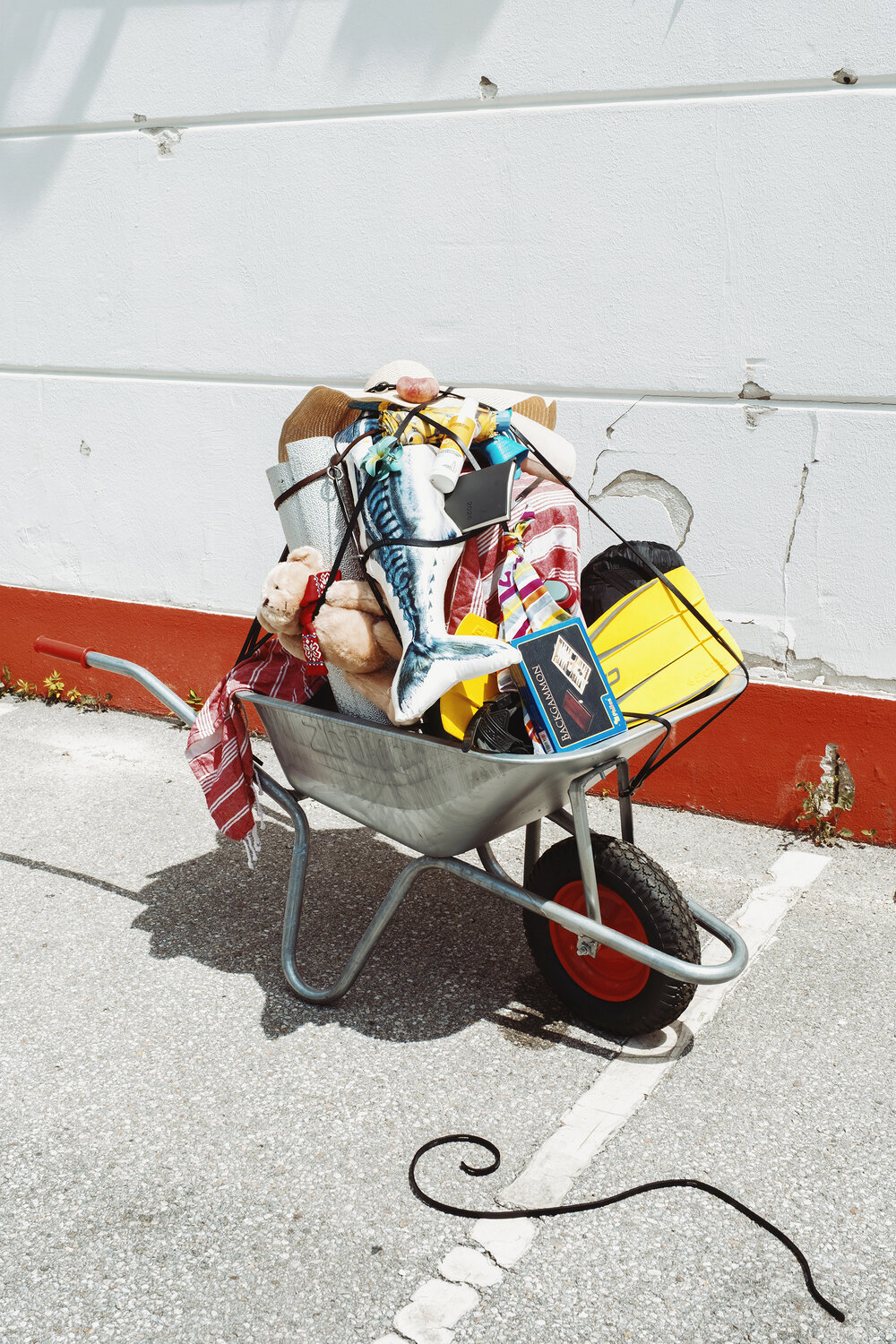 A metal wheelbarrow filled with various items—including a teddy bear, a yellow toolbox, a scarf, an INA KENT box, and more—stands on a paved surface near a white wall.