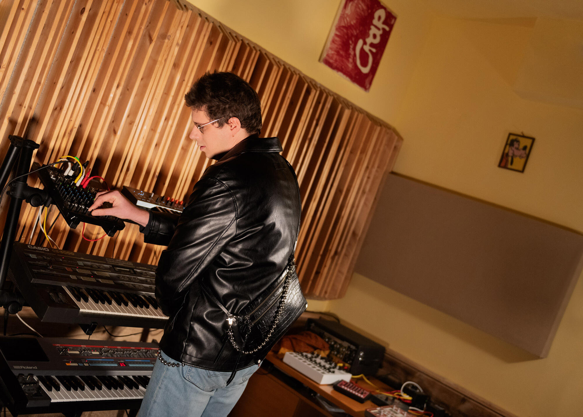 Eine Person steht in einem Musikstudio, trägt eine schwarze Lederjacke von INA KENT und bedient ein Synthesizer-Keyboard, umgeben von Audiogeräten vor dem Hintergrund einer hölzernen Akustikplatte.