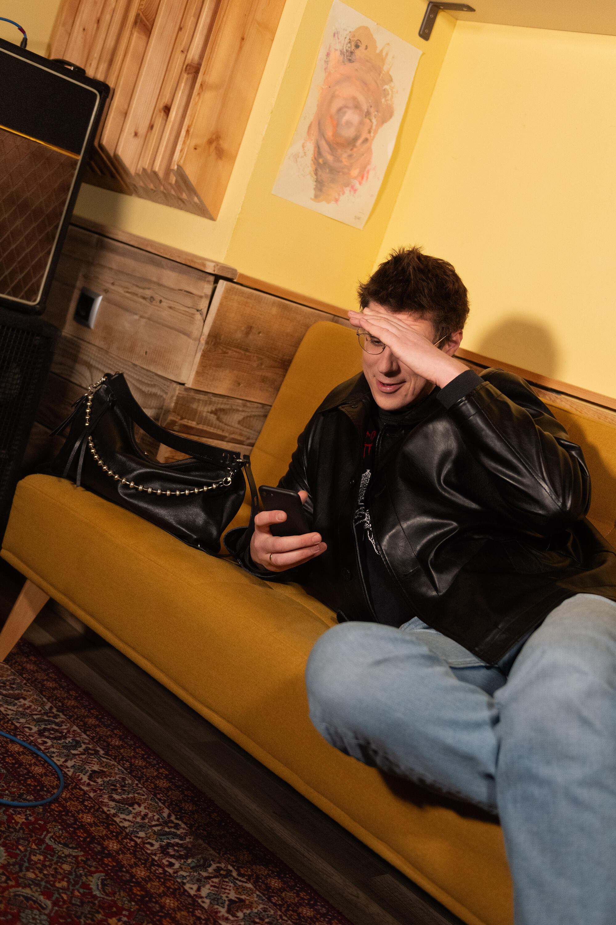 Eine Person in einer schwarzen Lederjacke sitzt auf einem gelben Sofa, schaut auf ihr Telefon und beschattet dabei ihre Augen mit einer Hand. Neben ihr steht eine Handtasche von INA KENT.