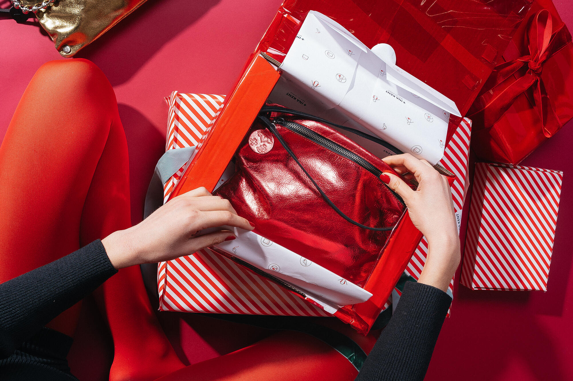 A person in red tights and a black top opens a red and white striped wrapping paper containing the MOONLIT ed.1 bag in the color Eco Antique Metallic Chili Red. The shiny, metallic red bag lies in a red gift box, surrounded by other red and white striped and red gift packages.