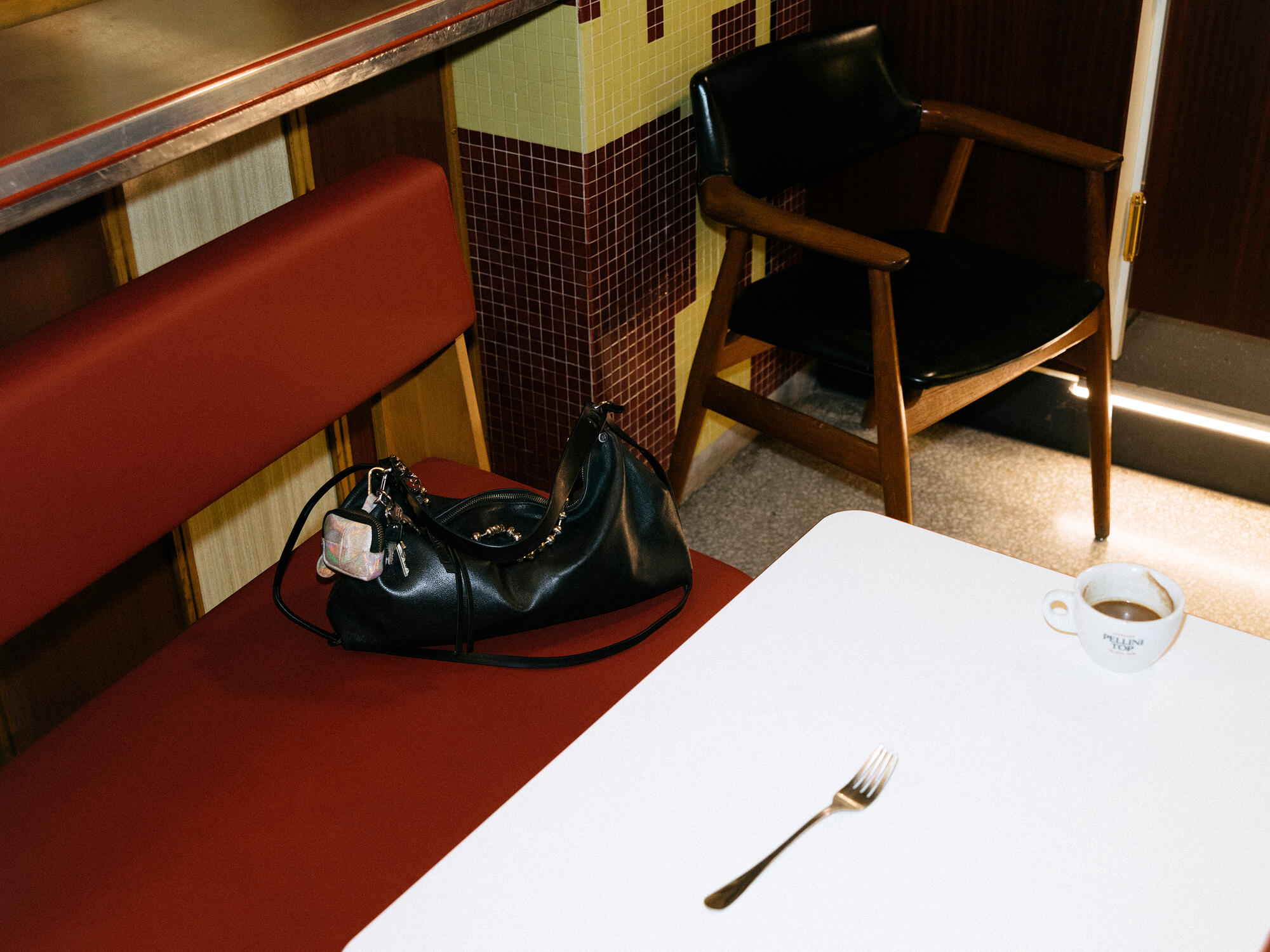 A black INA KENT bag on the red bench of a retro diner. Next to it, a white table with a single fork and a half-full espresso cup - as if someone had left their seat.