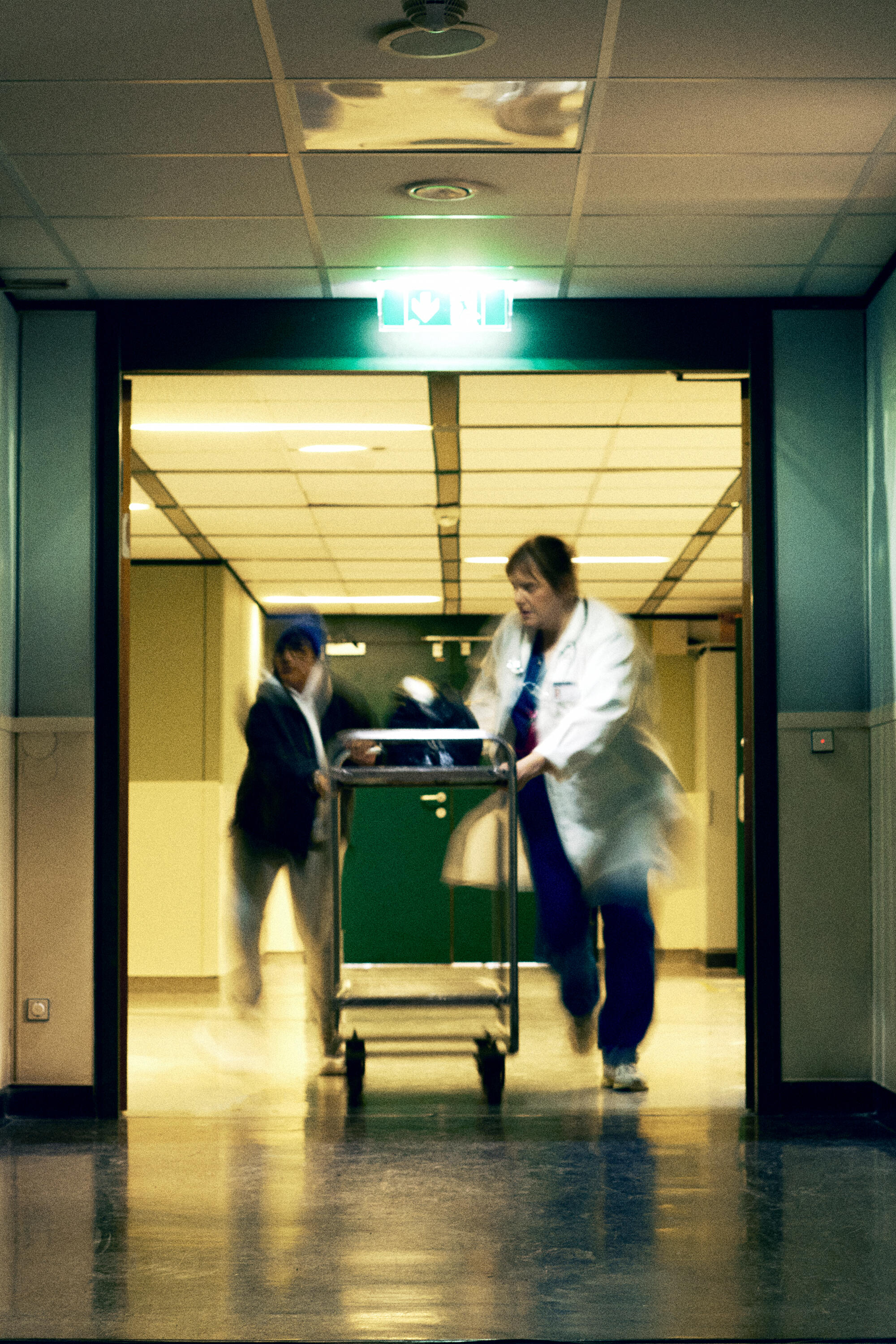 Zwei medizinische Fachkräfte transportieren einen Patienten auf einer Trage unter Neonlicht durch einen Krankenhausflur, während eine INA KENT-Tasche sicher neben einem der Ärzte liegt.