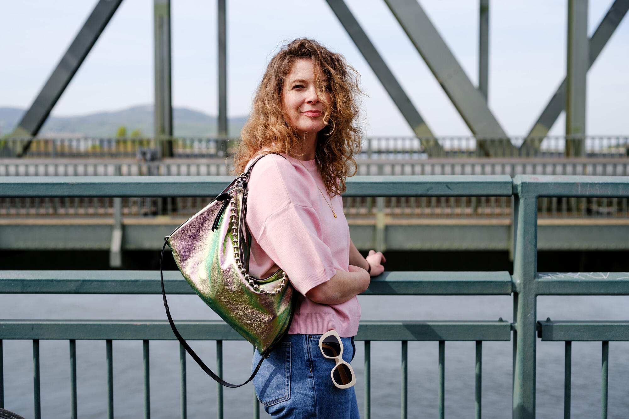 A woman with curly hair in a pink top and jeans stands by a metal railing on a bridge, carrying an iridescent INA KENT bag and holding sunglasses.