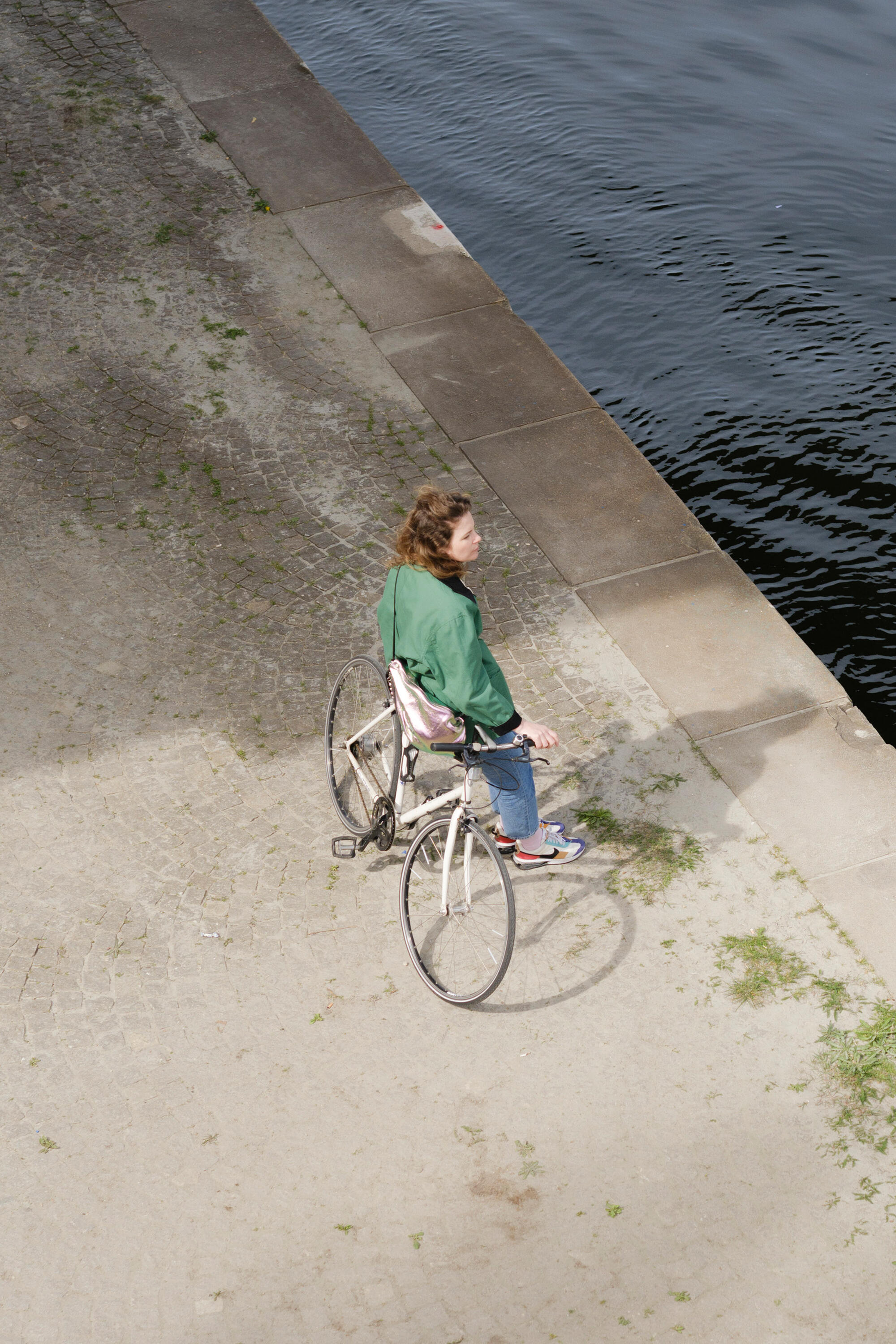 A person in a green jacket, carrying an INA KENT bag, sits on a bicycle near a paved path beside a body of water.