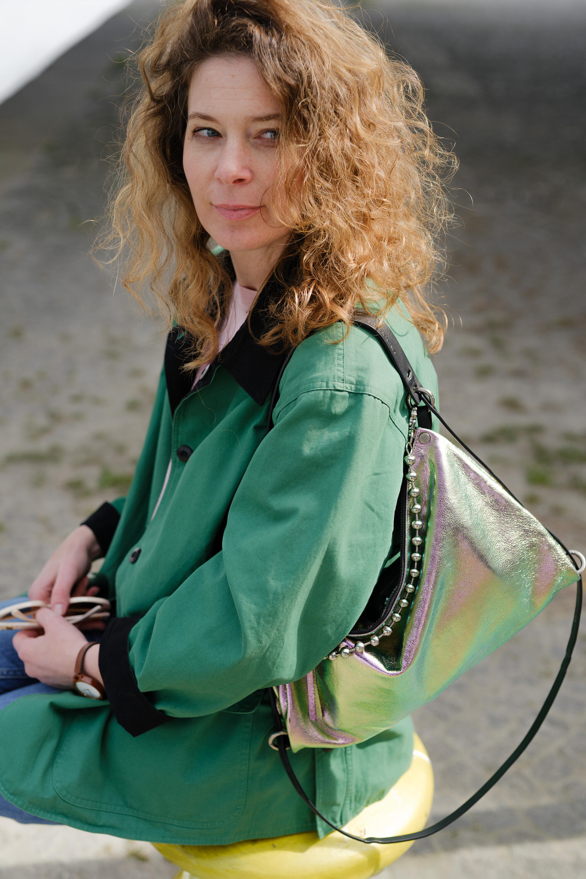 Woman with curly hair wearing a green coat sits outdoors, an INA KENT metallic iridescent bag on her shoulder, looking slightly to the side.