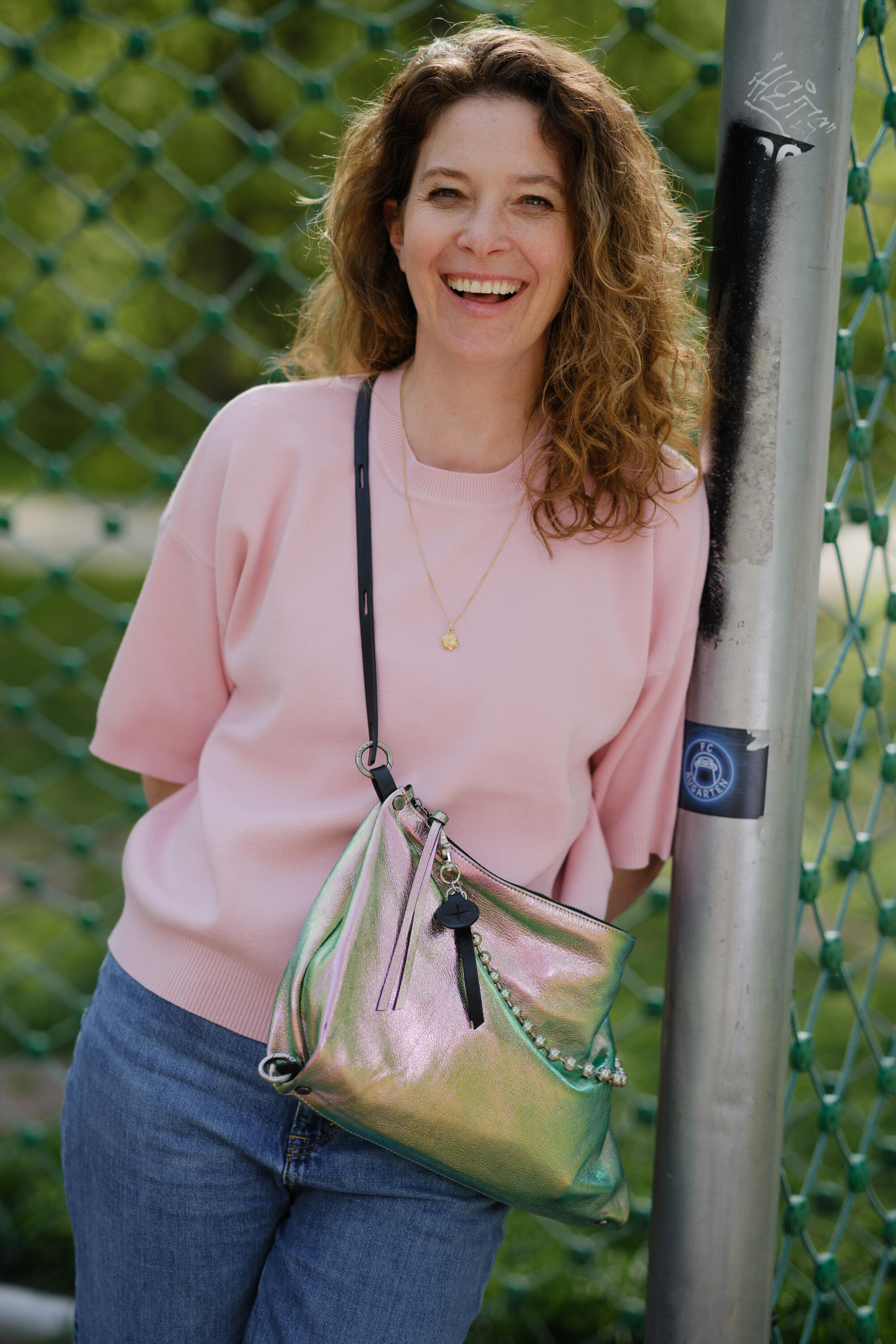 A woman with curly hair wearing a light pink sweater and blue jeans smiles while leaning against a metal pole, carrying a shiny iridescent INA KENT handbag.