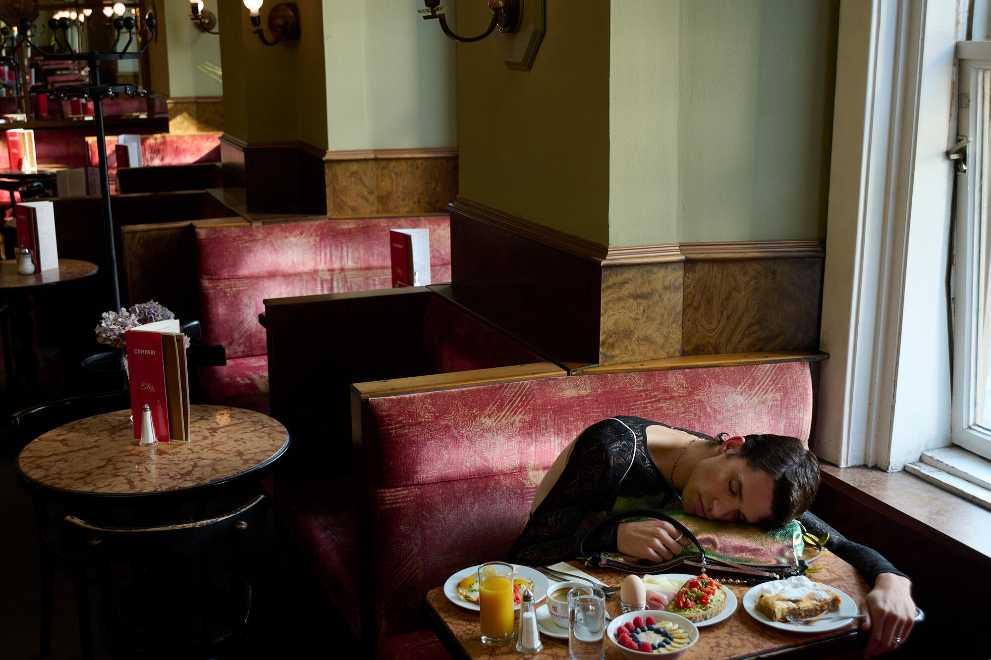 A person is asleep on a red booth in a restaurant, their head resting on the table next to a tray with breakfast food, drinks, and an INA KENT bag by their side.