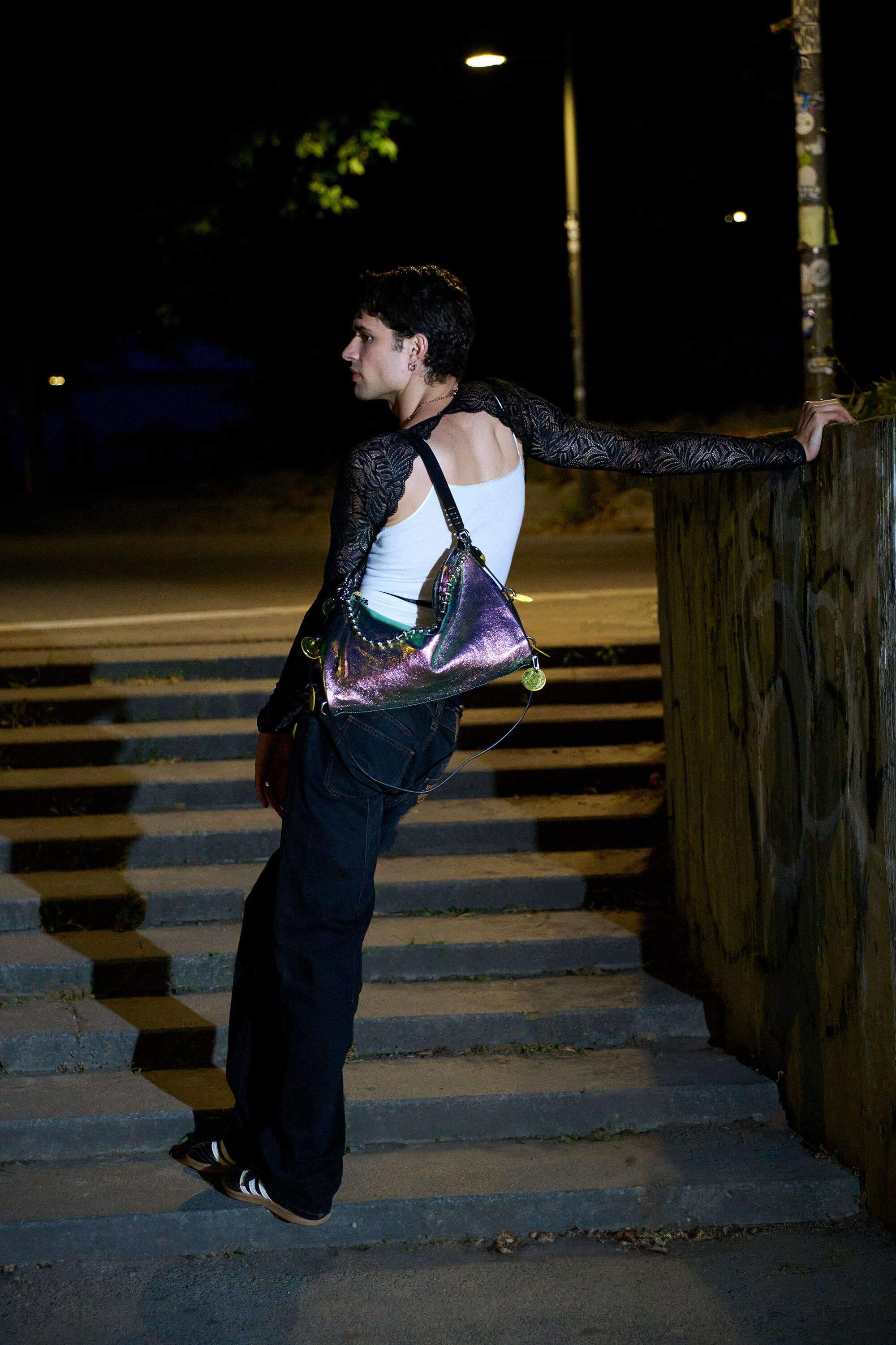 A person in black pants, a white tank top, and a lace long-sleeve leans on a wall by steps at night, carrying a shiny INA KENT iridescent handbag over their shoulder.