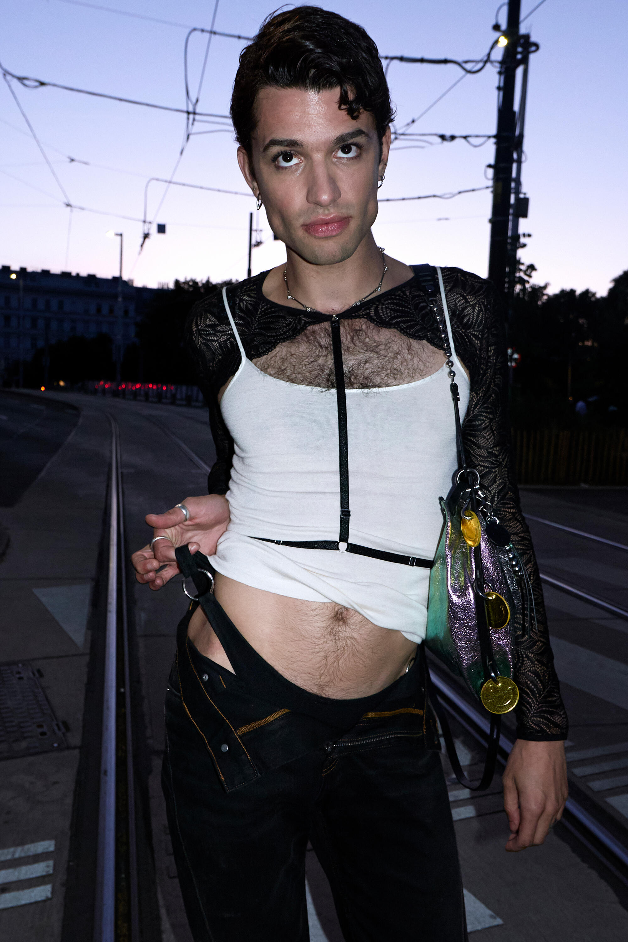 A person stands on tram tracks at dusk, wearing a white tank top over a black lace long-sleeve, unbuttoned pants, and holding an INA KENT metallic purse adorned with keychains.