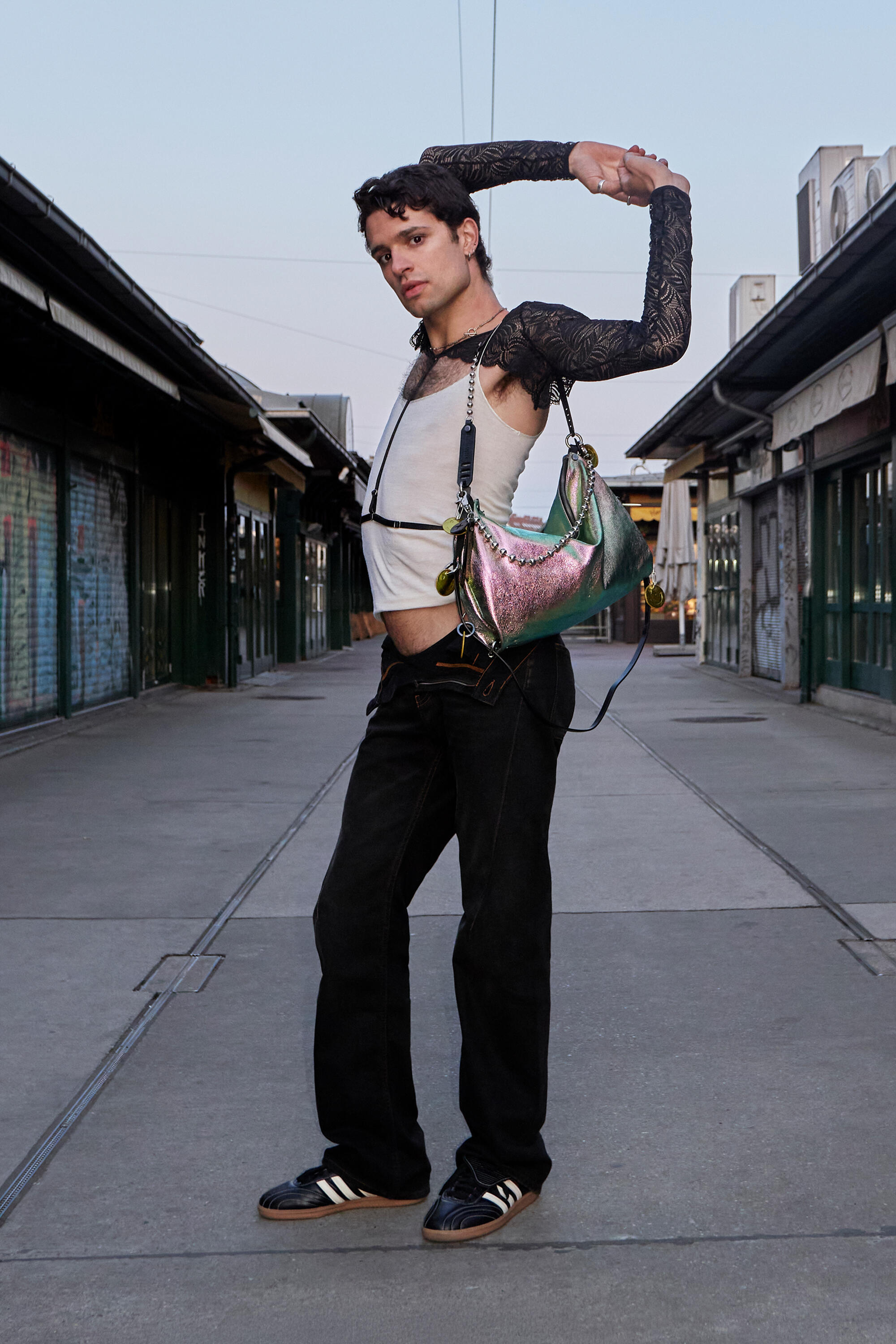 A person wearing a white tank top, black pants, and black sneakers poses in an empty outdoor walkway, holding a large INA KENT handbag with one arm raised over their head.