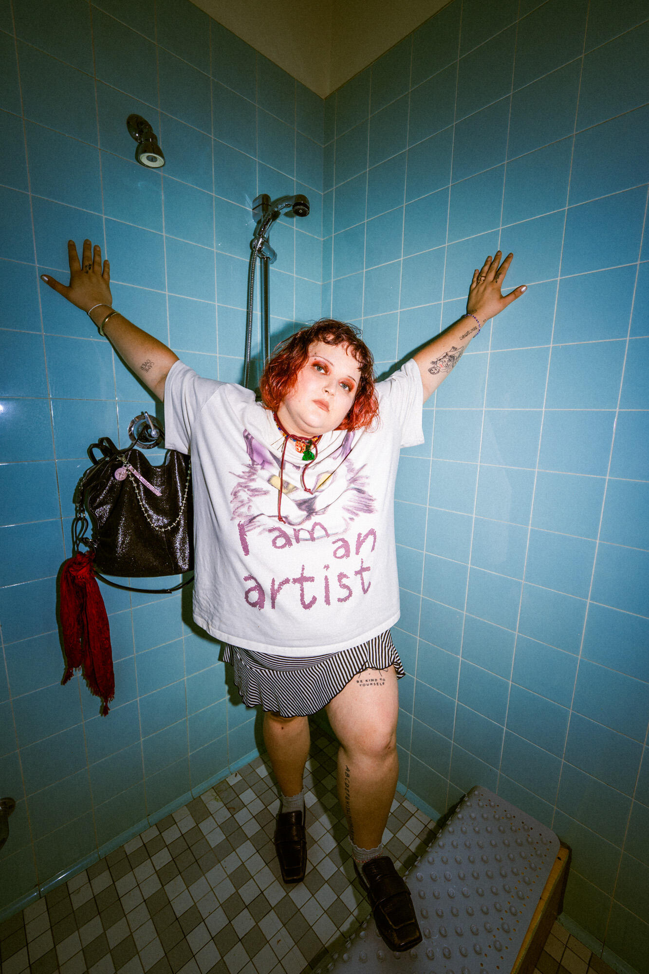 Christl Christiane Rechenmacher von mala kolumna steht in einer türkisfarbenen Duschkabine im Radiokulturhaus, trägt ein „I am an artist“-Shirt und hat die schwarze INA KENT Tasche DELA EDEN ed.1 neben sich hängen.