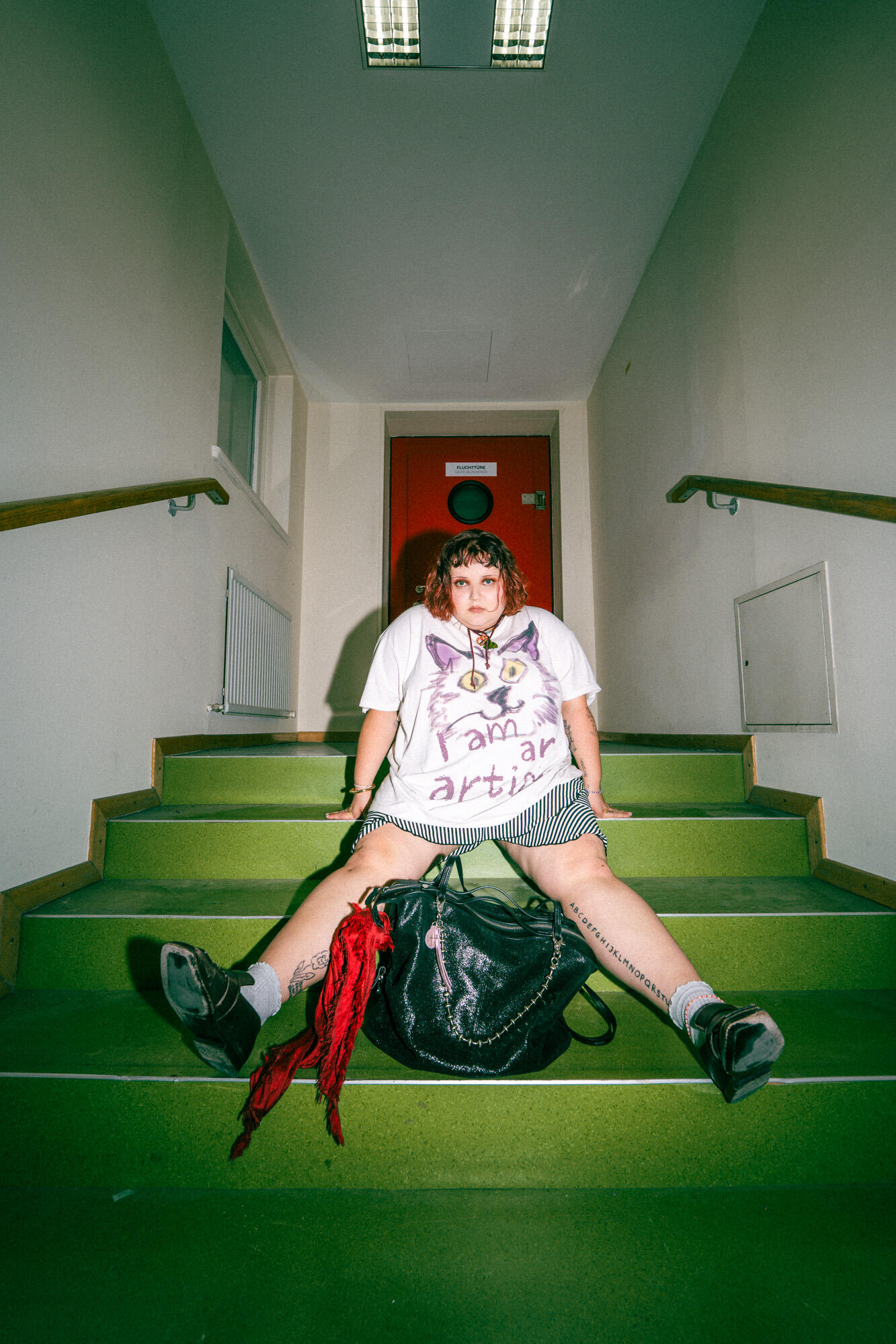 Person sitting on green stairs with legs spread, wearing a white graphic t-shirt, striped shorts, and black shoes, with an INA KENT black backpack and red scarf in front of them.
