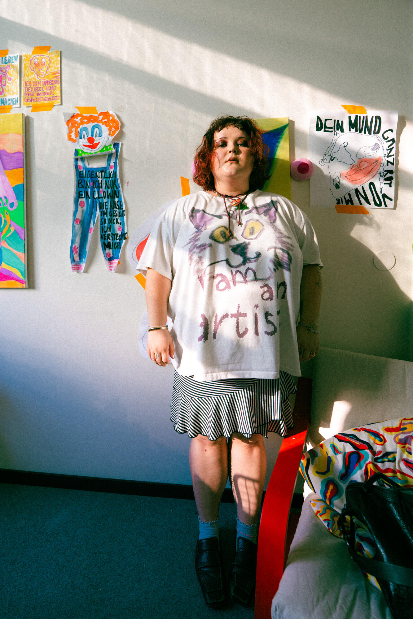 Christl Christiane Rechenmacher steht in ihrem Atelier vor bunt gestalteten Kunstwerken an der Wand, trägt ein „I am an artist“-Shirt und blickt ruhig in die Kamera.