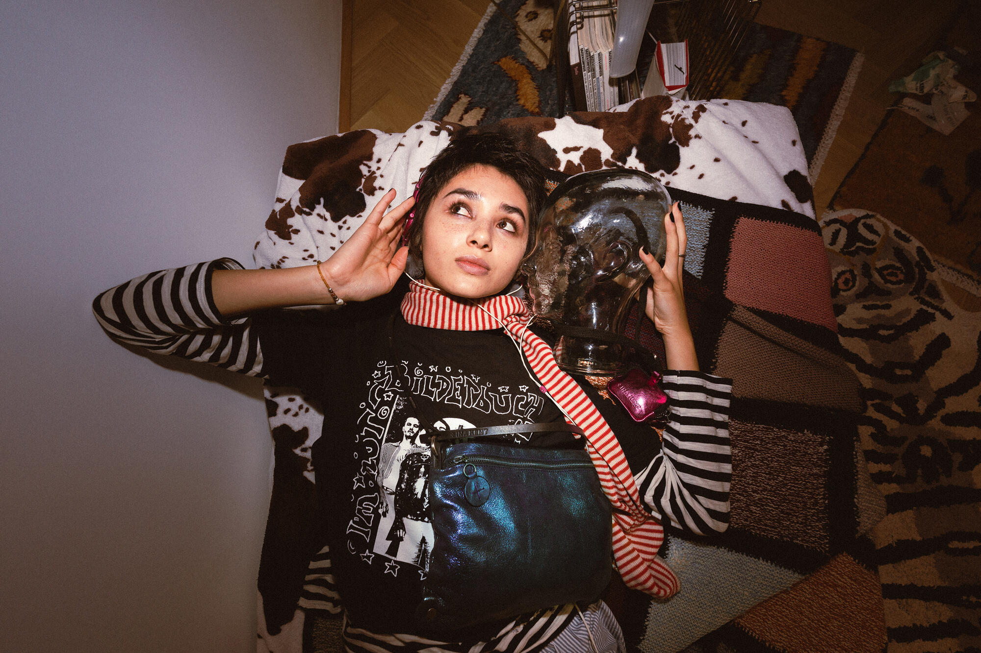 A person lies on a patterned rug holding a glass head, wearing a striped shirt and red scarf, with an INA KENT bag resting on their chest.