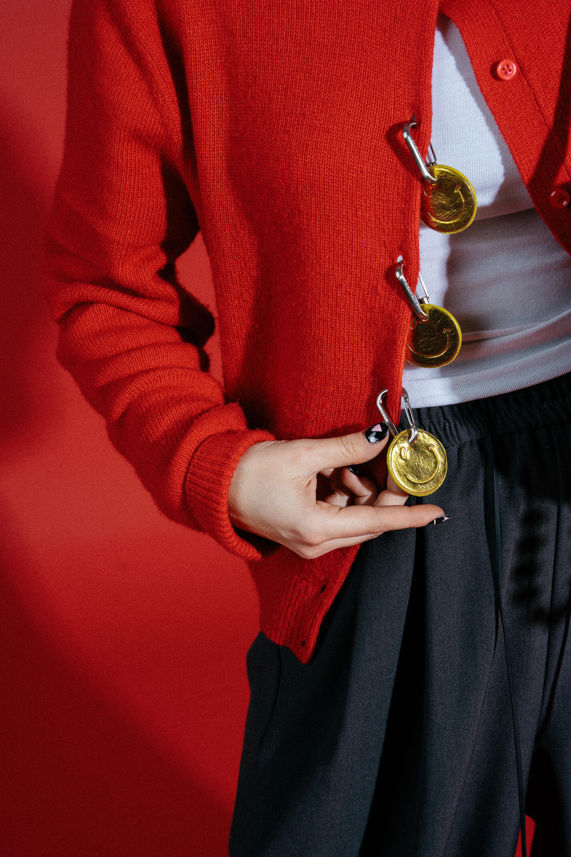 Eine Person, die eine rote Strickjacke mit großen Sicherheitsnadeln und goldenen Medaillen über einem weißen Hemd und einer grauen Hose trägt, steht vor einem roten Hintergrund und ist mit einer stilvollen INA KENT-Tasche ausgestattet.