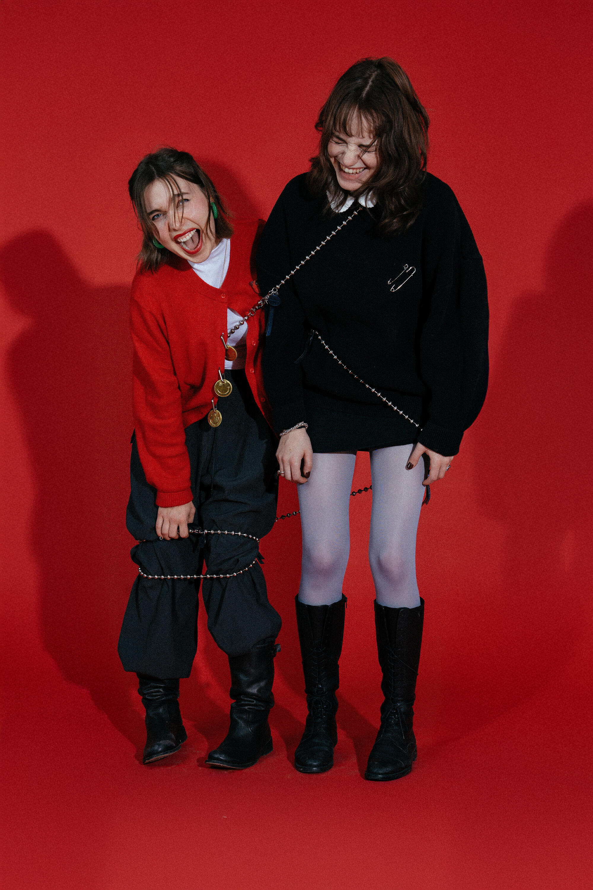 Zwei junge Frauen, die eine in einem roten Pullover, die andere in Schwarz mit weißer Strumpfhose, stehen lachend vor einem kräftigen roten Hintergrund. Sie sind durch eine INA KENT-Kette miteinander verbunden, was ihre spielerische Verbindung unterstreicht.