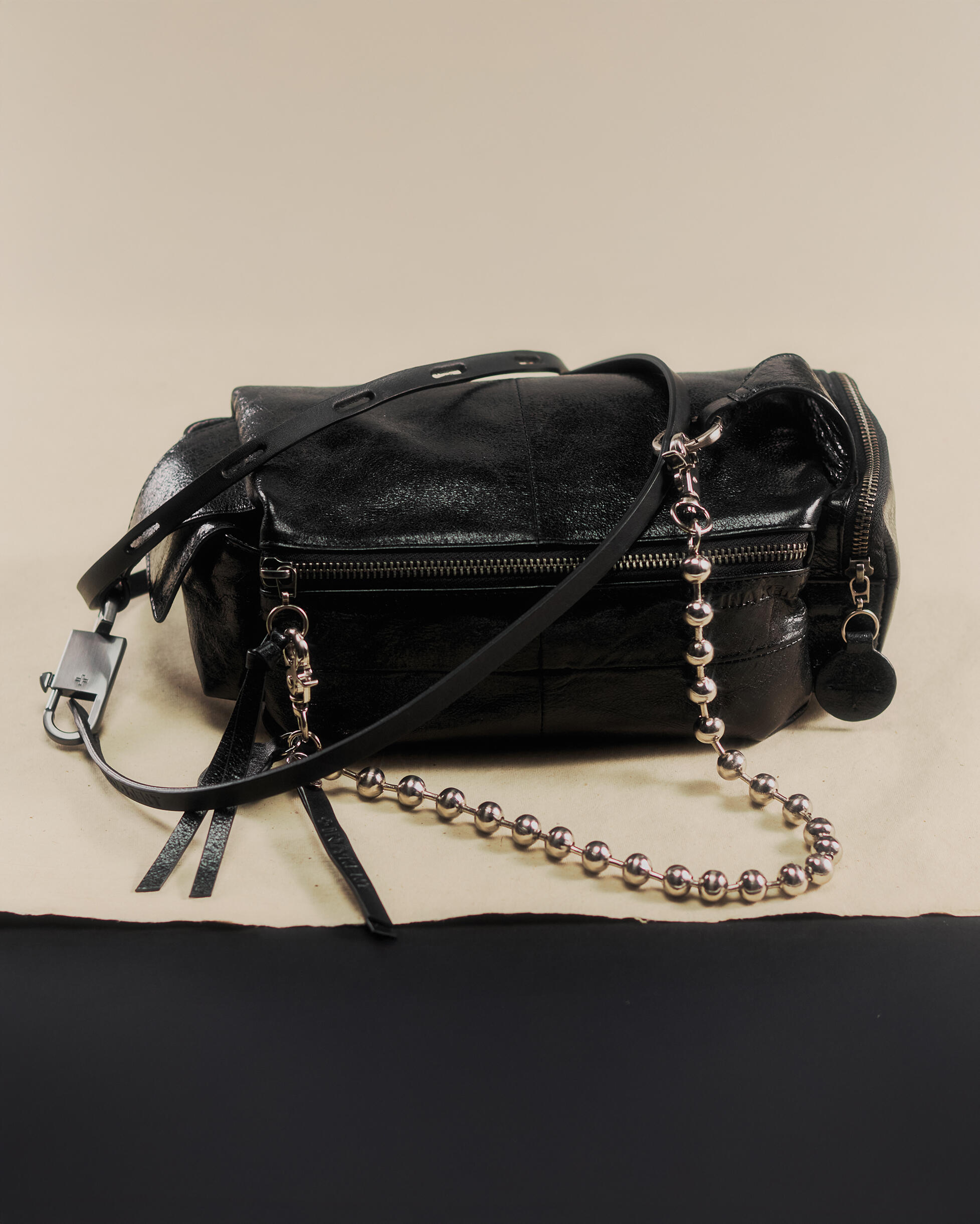 Eine schwarze Lederhandtasche von INA KENT mit silbernen Beschlägen, zwei Reißverschlüssen und einem dicken Kettenriemen wird auf einer beige-schwarzen Fläche ausgestellt.