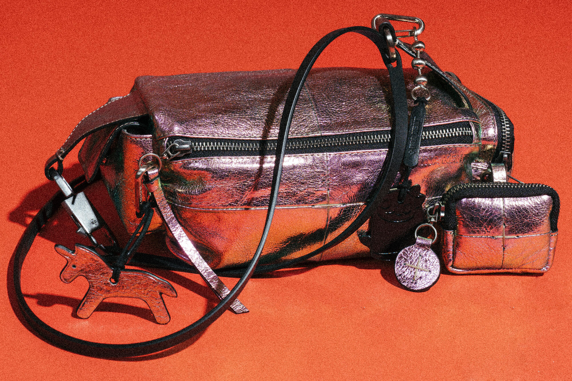 Eine metallisch lilafarbene INA KENT Handtasche mit passendem Münzbeutel, schwarzem Riemen und hundeförmigem Schlüsselanhänger auf rotem Hintergrund.