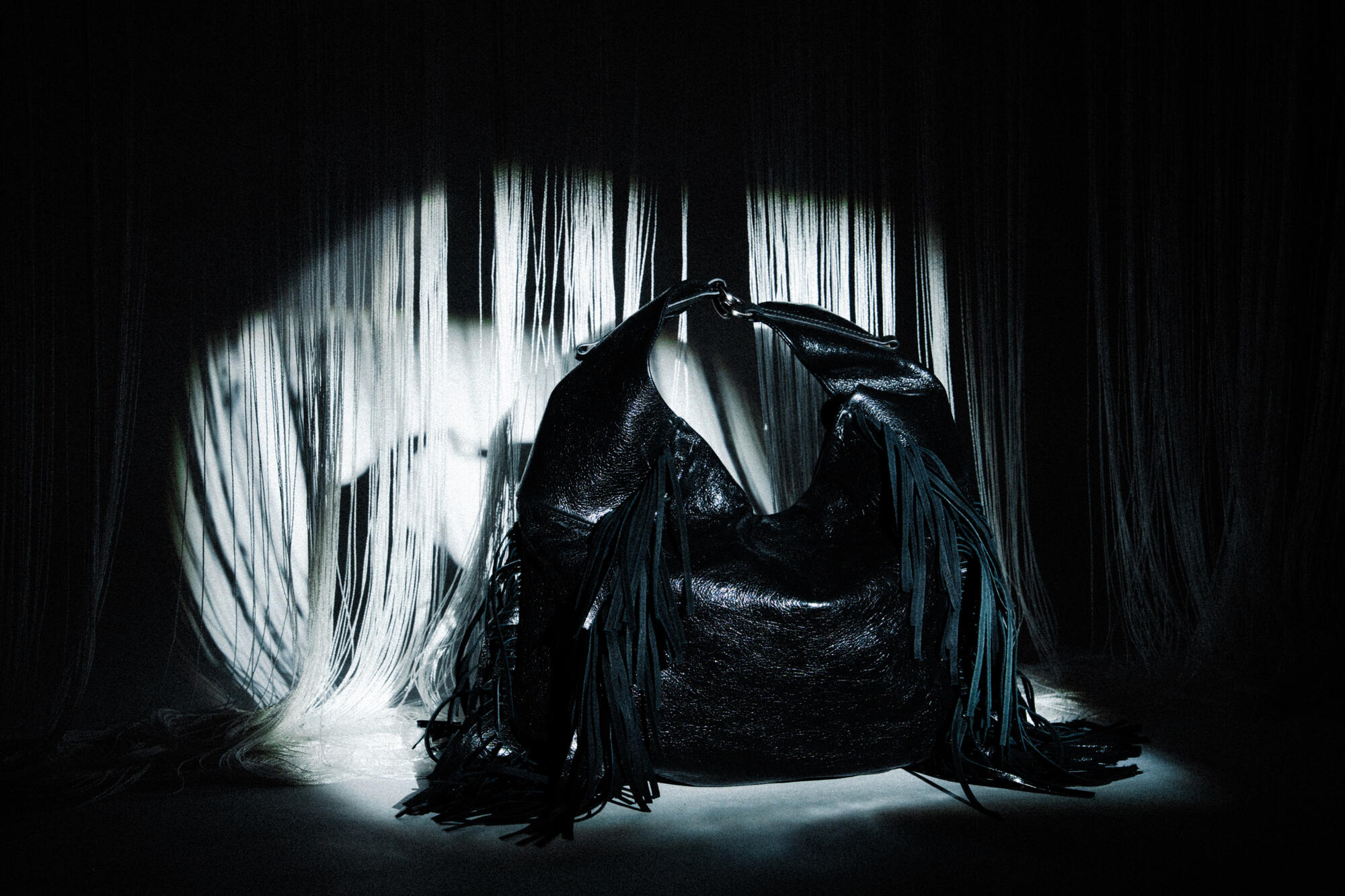 An INA KENT black slouchy bag with fringe details is displayed under dramatic lighting, with draped fabric in the background.