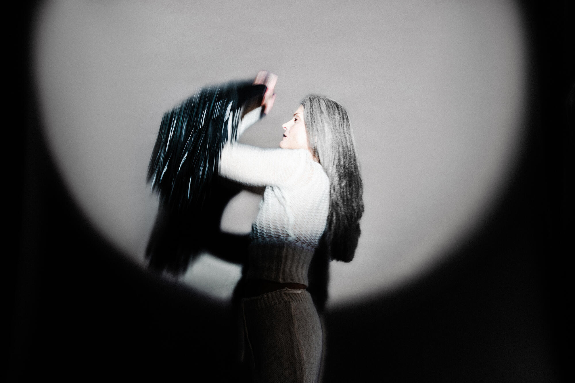 A person with long hair stands in a spotlight, wearing a light sweater and holding up an INA KENT dark, sparkly garment in motion.