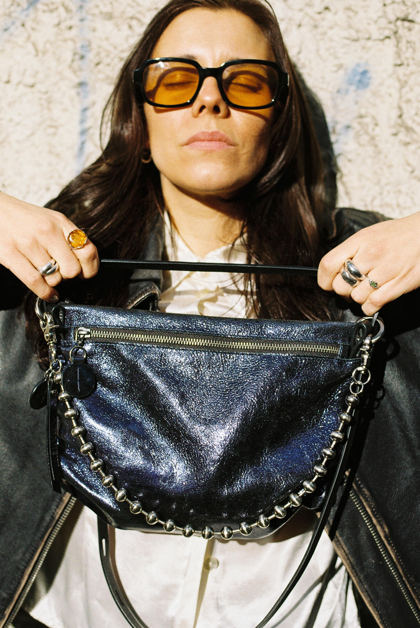 Eine Person, die eine Sonnenbrille und eine Lederjacke trägt, hält eine glänzende schwarze Handtasche von INA KENT mit Kettenverzierung vor sich an eine strukturierte Wand.