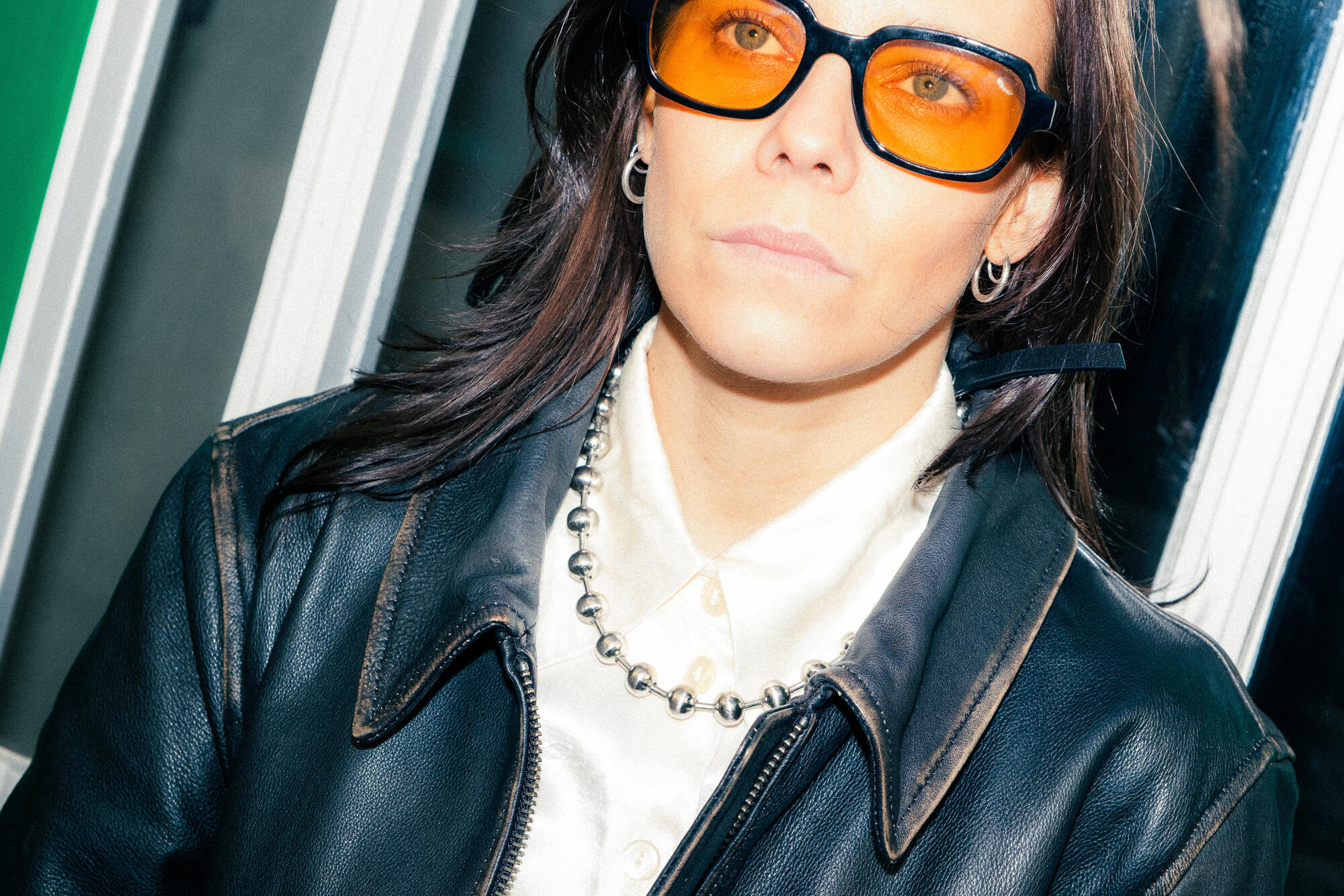 Person, die eine orangefarbene Brille, eine schwarze Lederjacke, ein weißes Hemd und eine silberne Perlenkette trägt und vor einem Schaufenster steht, wobei ein Accessoire von INA KENT den Look vervollständigt.