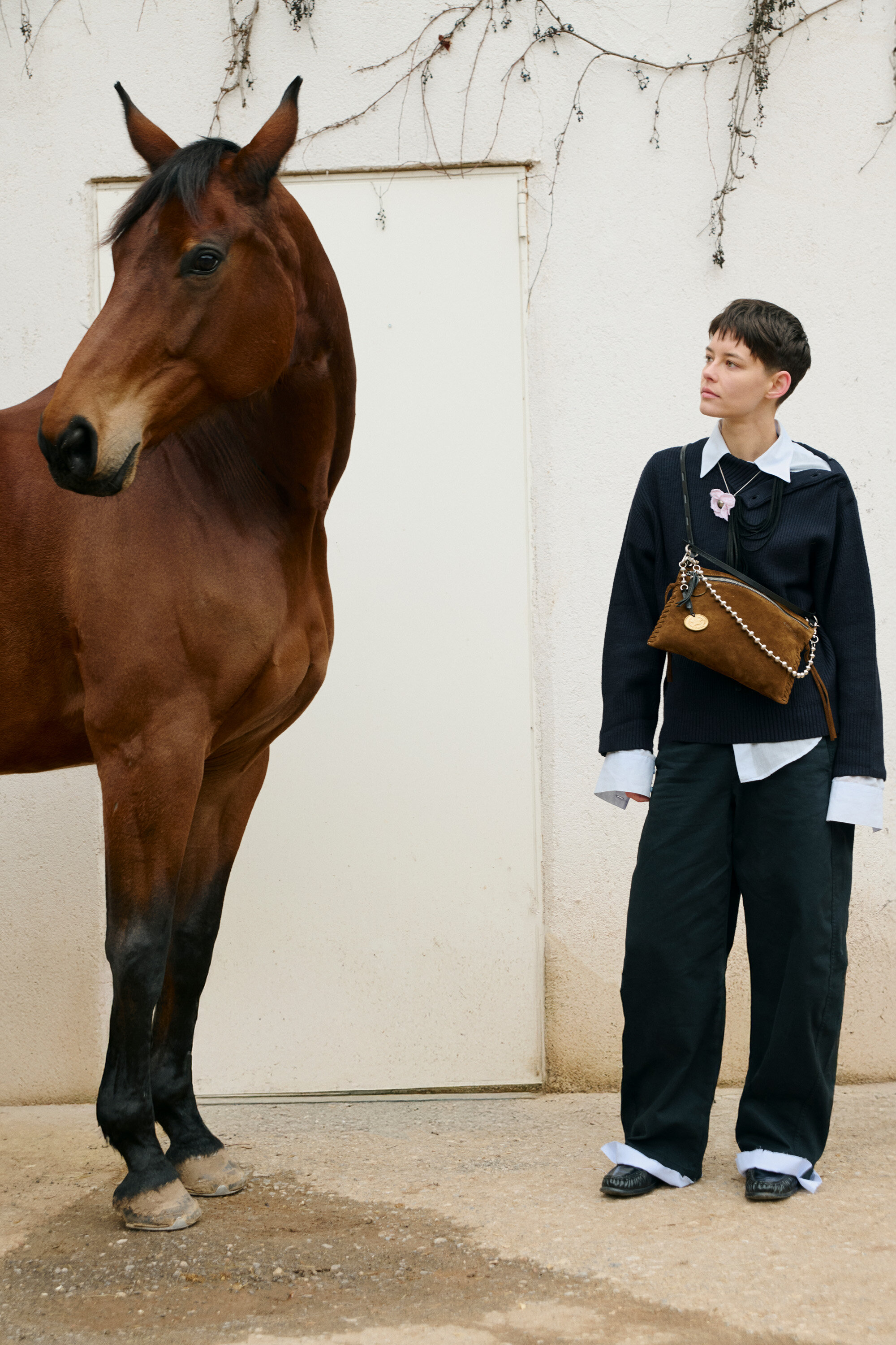A person wearing loose black pants, a black sweater, and an INA KENT brown bag stands next to a brown horse in front of a white wall with a closed door.