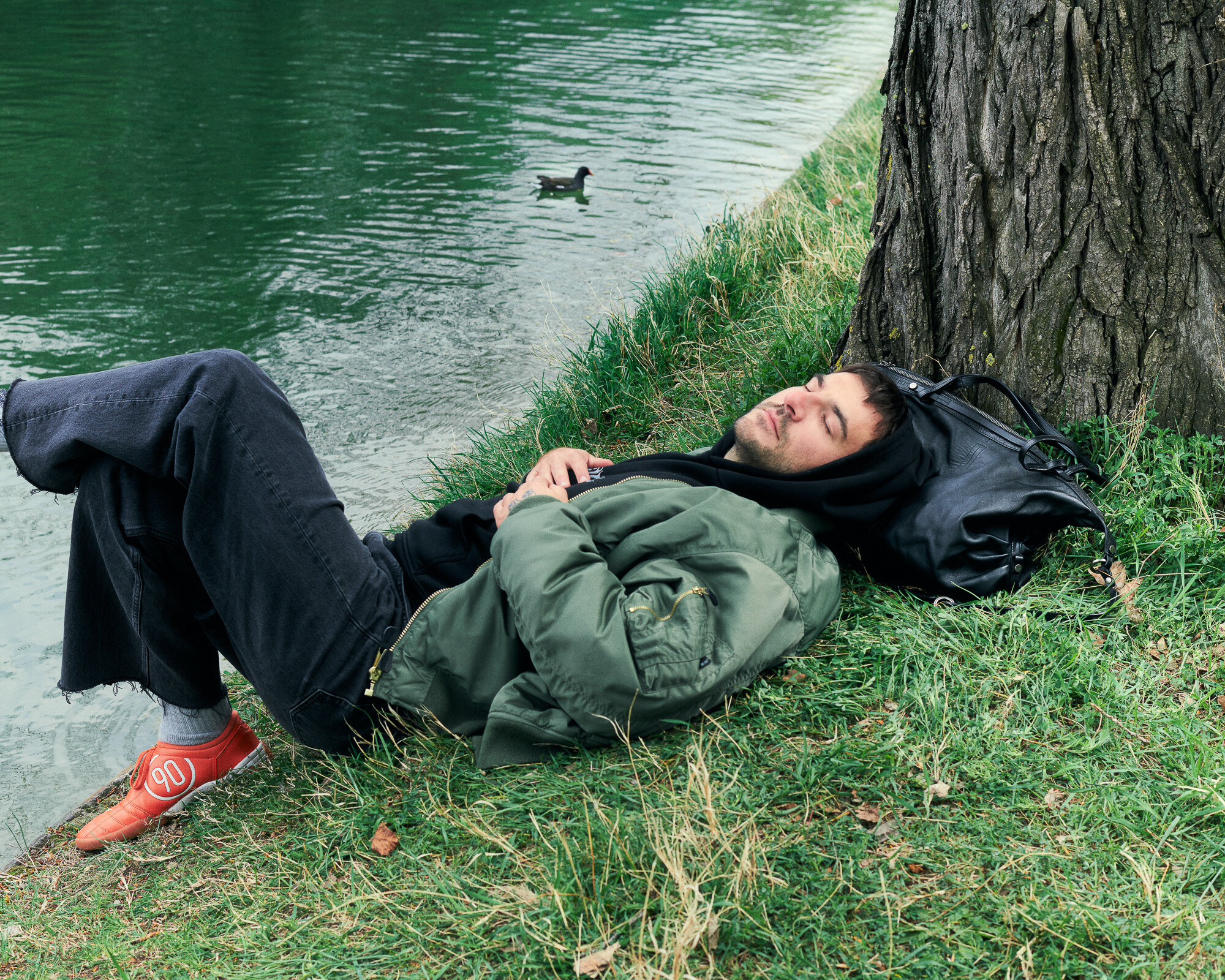 Eine Person mit grüner Jacke und orangefarbenen Schuhen liegt im Gras neben einem Baum und einem Gewässer und benutzt einen INA KENT-Rucksack als Kopfkissen. Eine Ente schwimmt in der Nähe im Wasser.