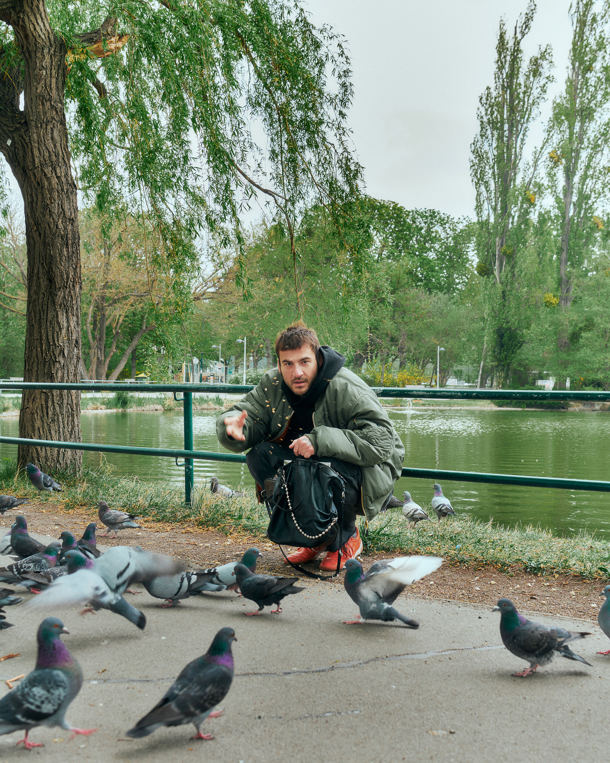 Ein Mann in grüner Jacke hockt auf einem Gehweg an einem Teich, umgeben von Tauben, und hält mit einer Hand Essen in die Höhe. Eine INA KENT-Tasche liegt an seiner Seite, im Hintergrund sind Bäume und Wasser zu sehen.