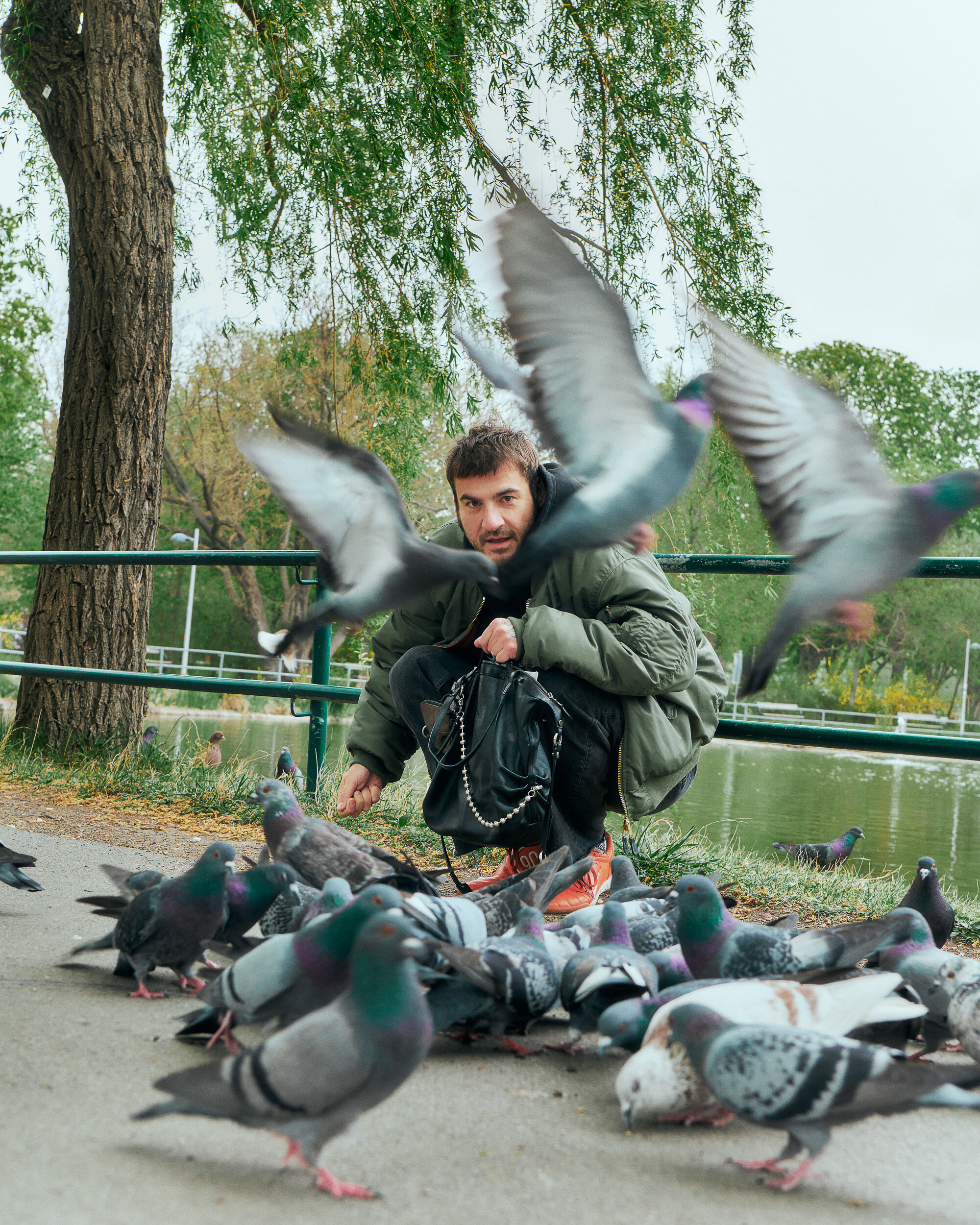 Ein Mann hockt an einem Teich und füttert eine Gruppe von Tauben, wobei mehrere Vögel um ihn herumfliegen und Bäume im Hintergrund zu sehen sind. Seine INA KENT-Tasche liegt in der Nähe.