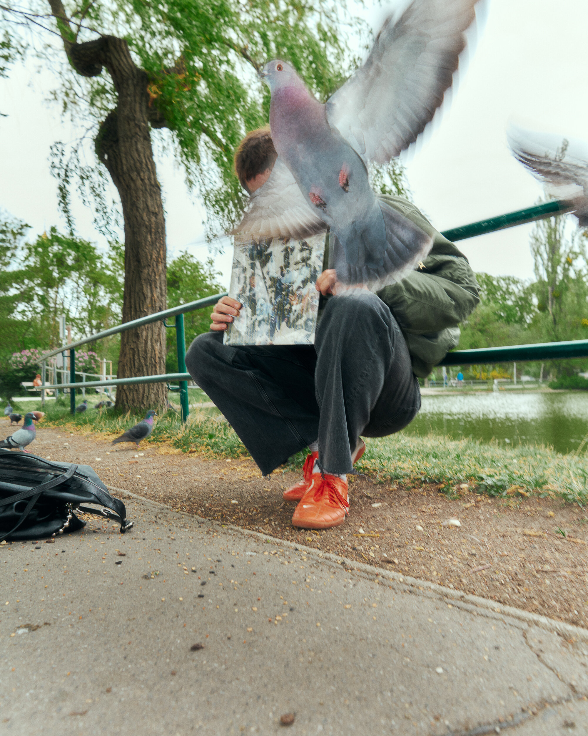 Person, die in der Nähe eines Teiches kauert und ein Magazin in der Hand hält, teilweise verdeckt von einer fliegenden Taube; ein INA KENT-Rucksack liegt in der Nähe auf dem Boden.