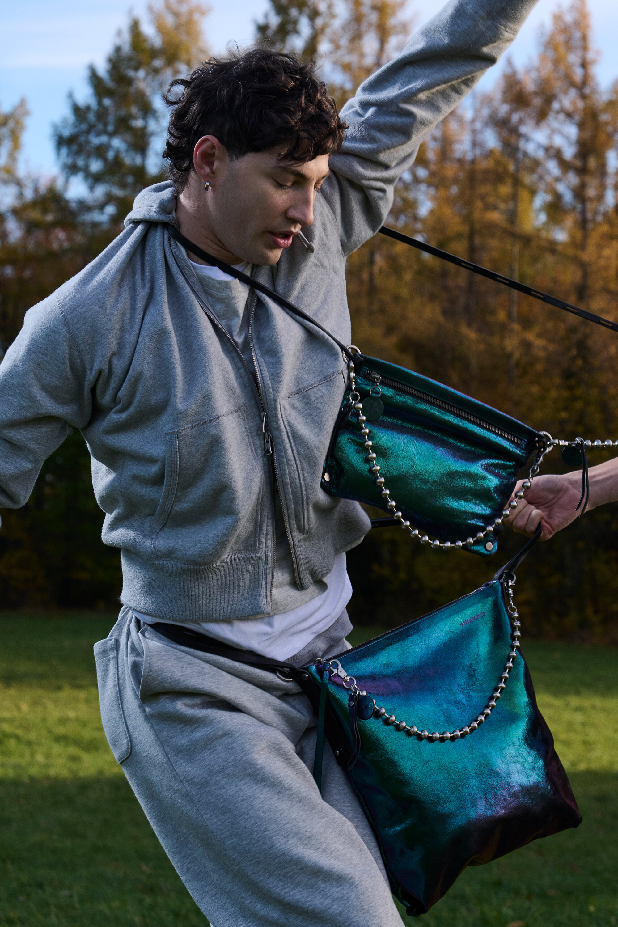 A person in a grey tracksuit poses outdoors with two iridescent INA KENT handbags, one worn across the chest and one at the waist, against a background of trees.