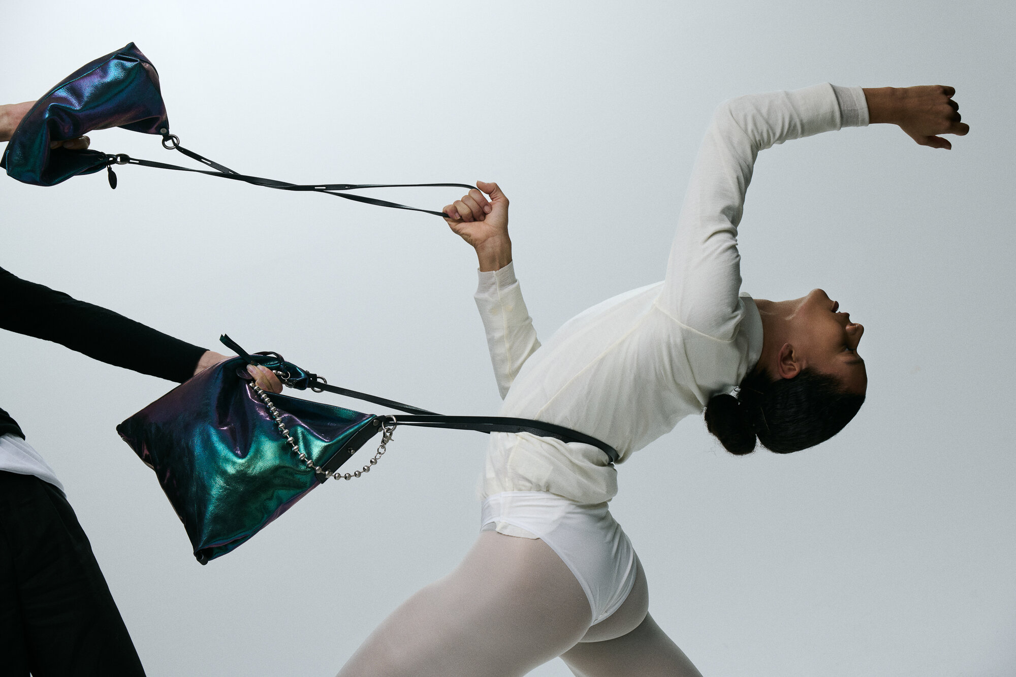 Eine weiß gekleidete Person lehnt sich dramatisch zurück und hält sich an den Riemen zweier schillernder INA KENT-Handtaschen fest, die von einer anderen schwarz gekleideten Person gezogen werden.