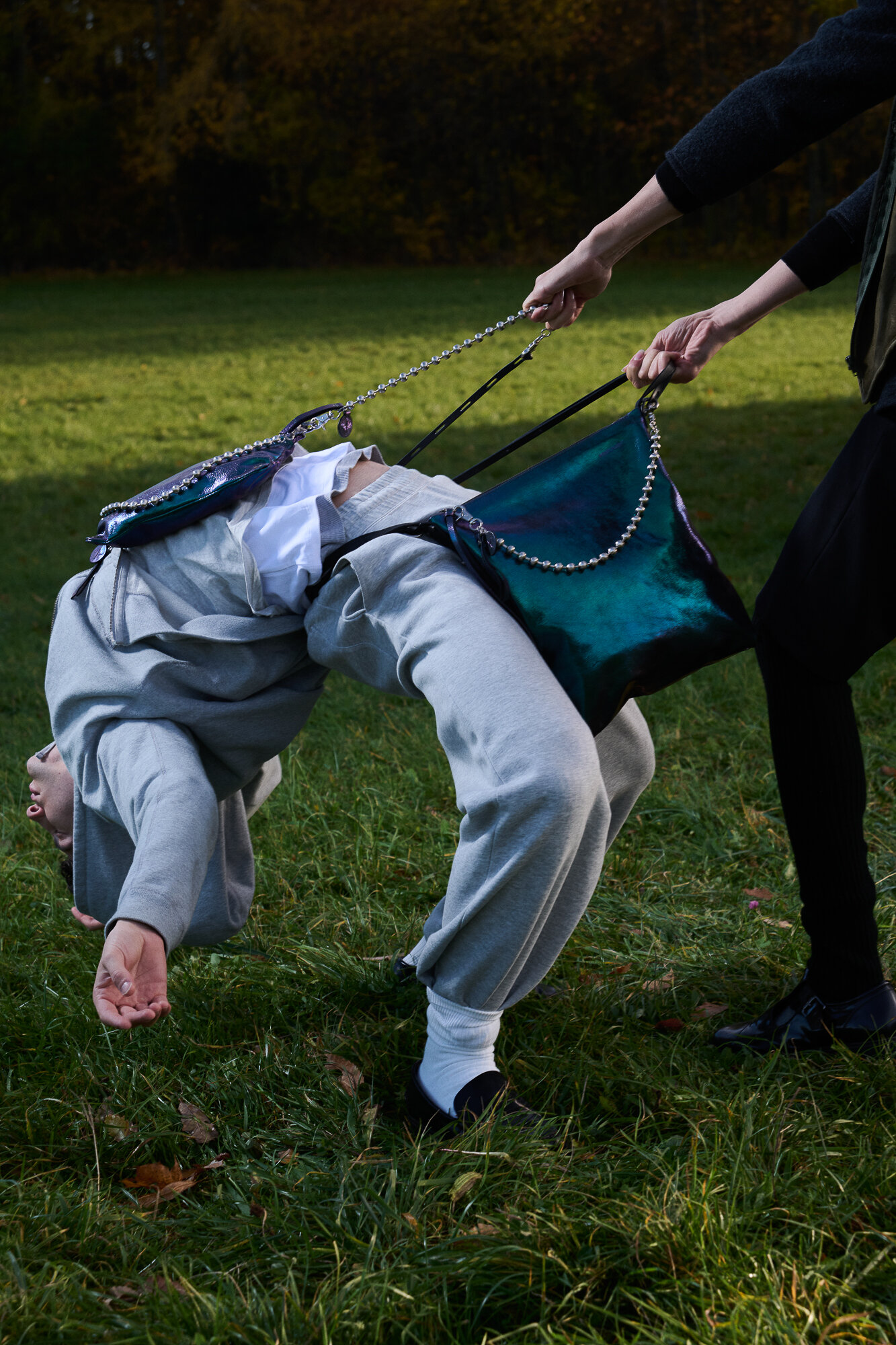 A person in grey sweats bends backwards while being held up by a large INA KENT green handbag with chain straps in an outdoor grassy area.