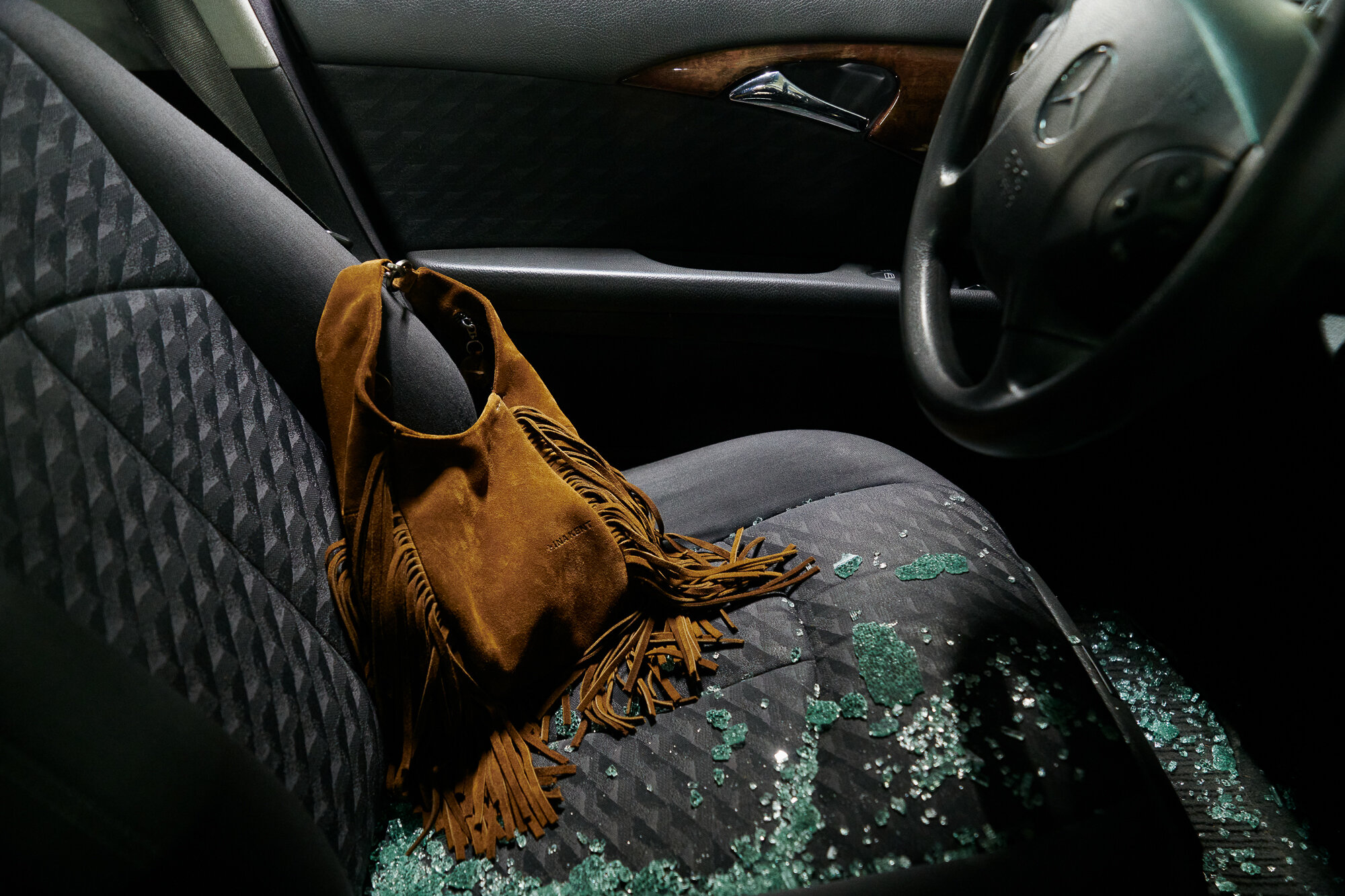 Eine braune Wildlederhandtasche mit Fransen von INA KENT liegt auf einem mit Glasscherben bedeckten Autositz neben dem Lenkrad, was auf einen kürzlichen Einbruch schließen lässt.