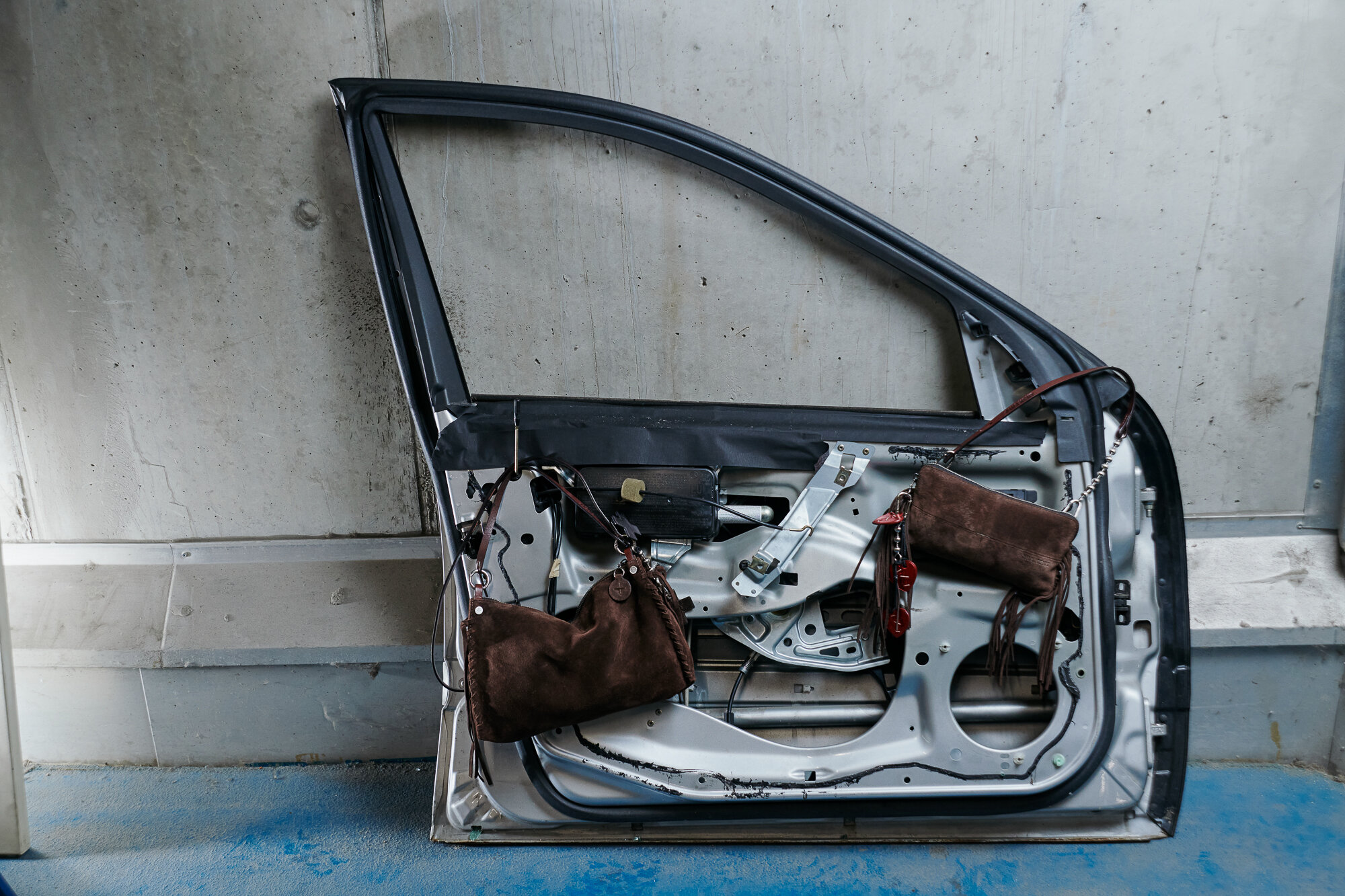 Ein Autotürrahmen lehnt an einer Betonwand, an dessen freiliegender Innenseite zwei braune INA KENT-Handtaschen hängen.
