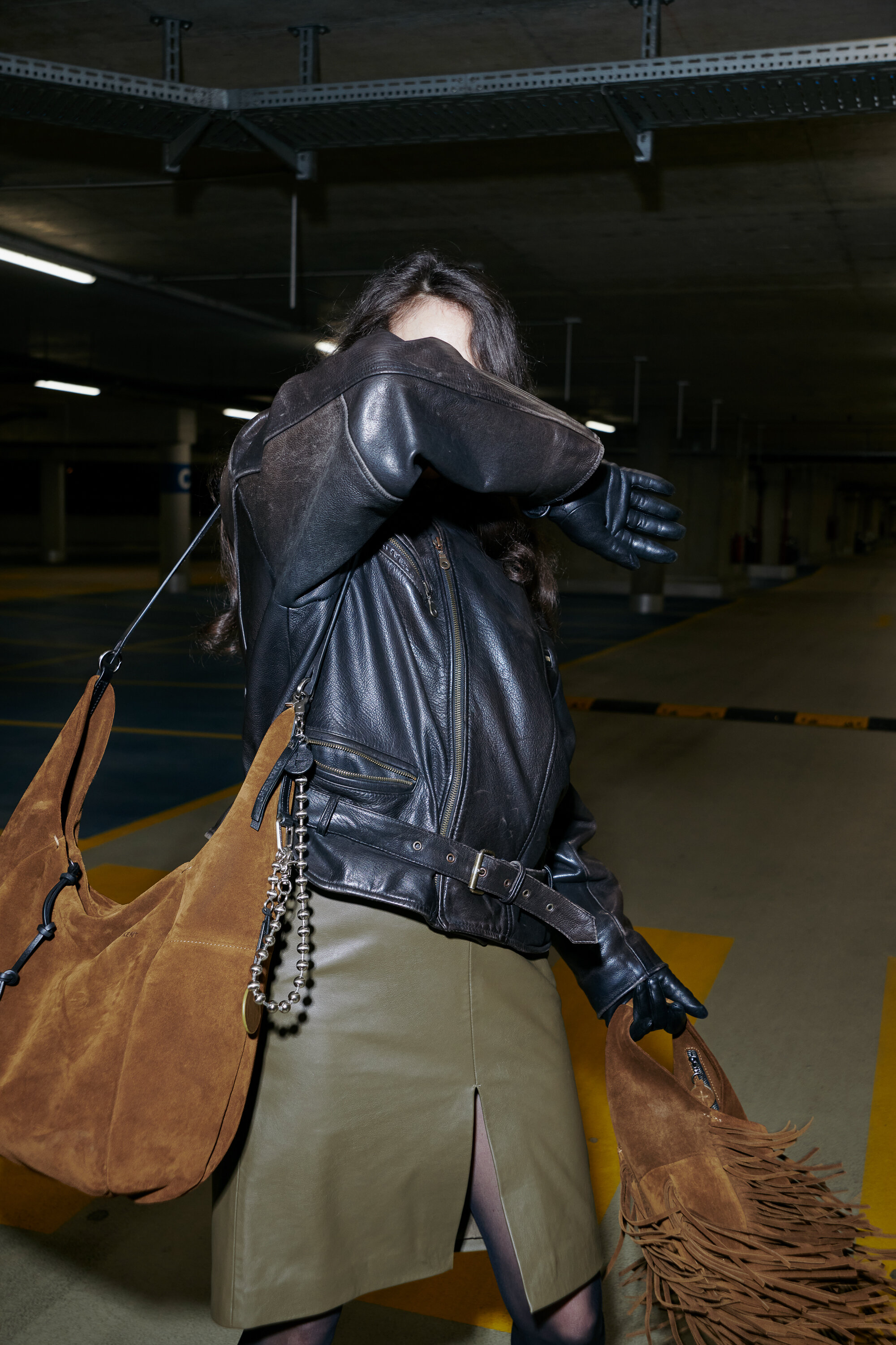 Eine Person in Lederjacke und Handschuhen hebt den Arm, um ihr Gesicht teilweise zu verdecken, und steht in einem leeren Parkhaus mit einer braunen Wildledertasche von INA KENT über der Schulter.