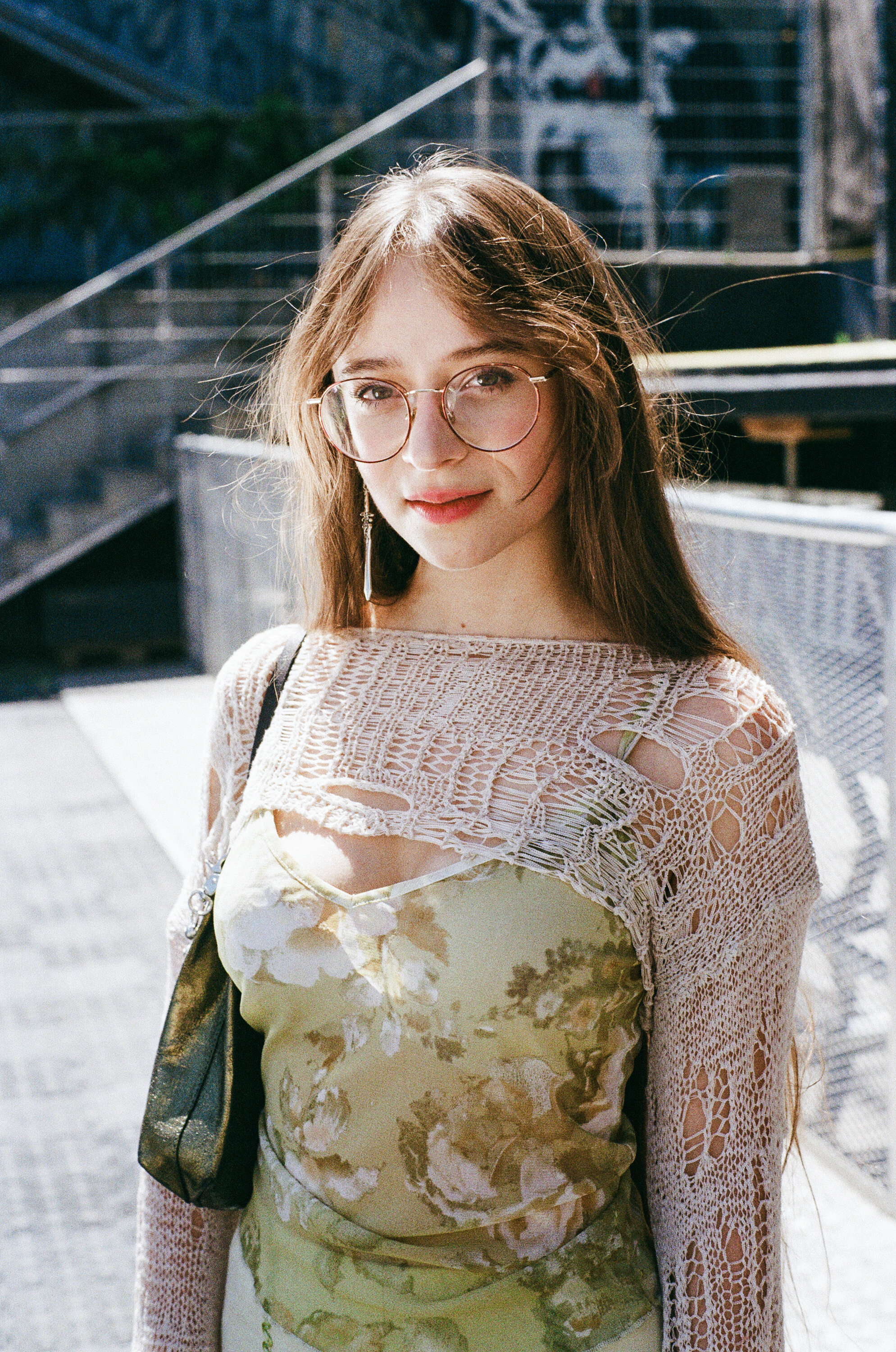 A young woman wearing glasses and a floral dress with a crochet top stands outdoors, looking at the camera. Metal stairs and railings are visible in the background, and she carries an elegant INA KENT bag.