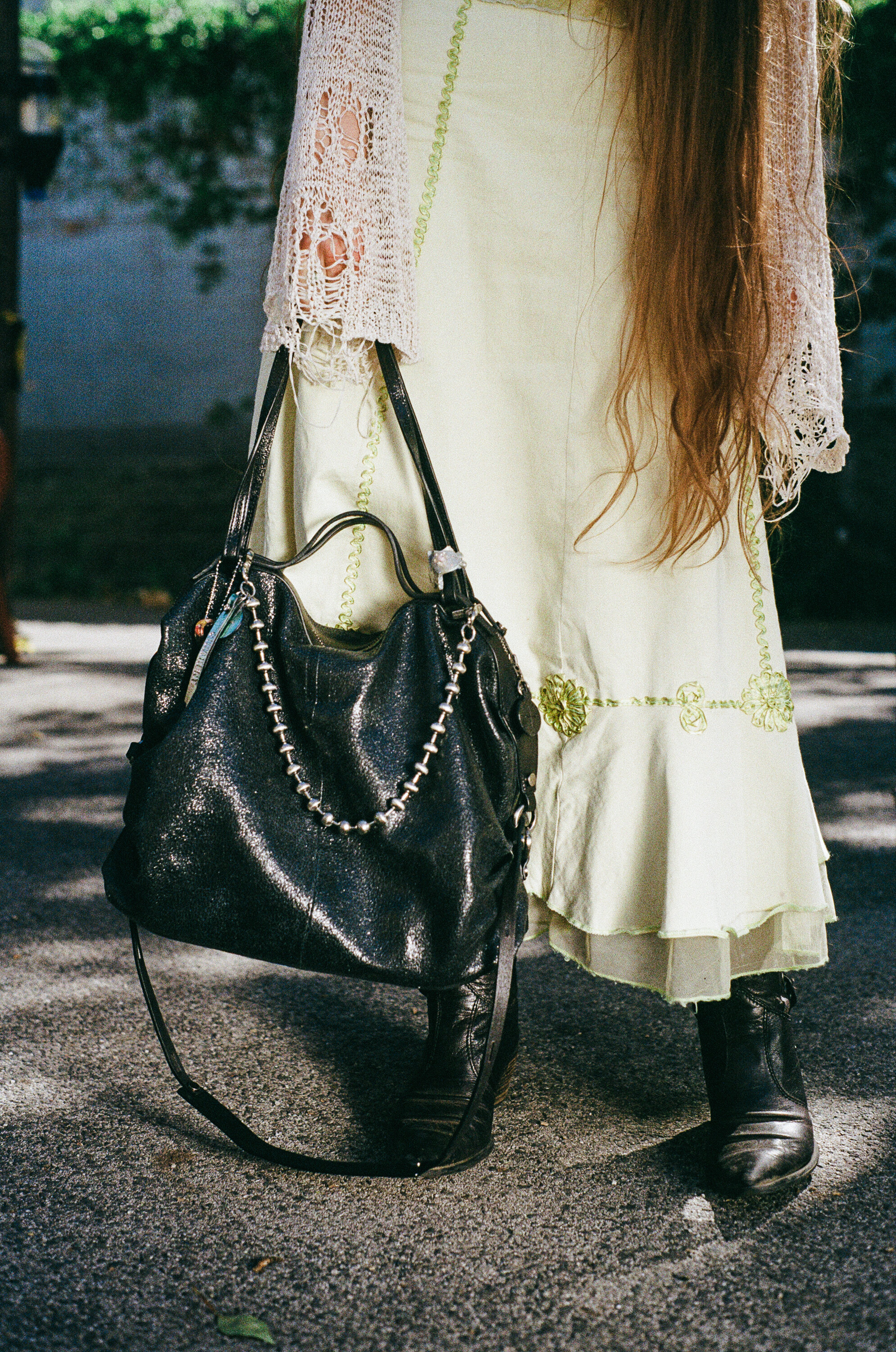 Person in a light green skirt and lace shawl standing outdoors, holding a large black INA KENT handbag with chain detail, wearing black boots.