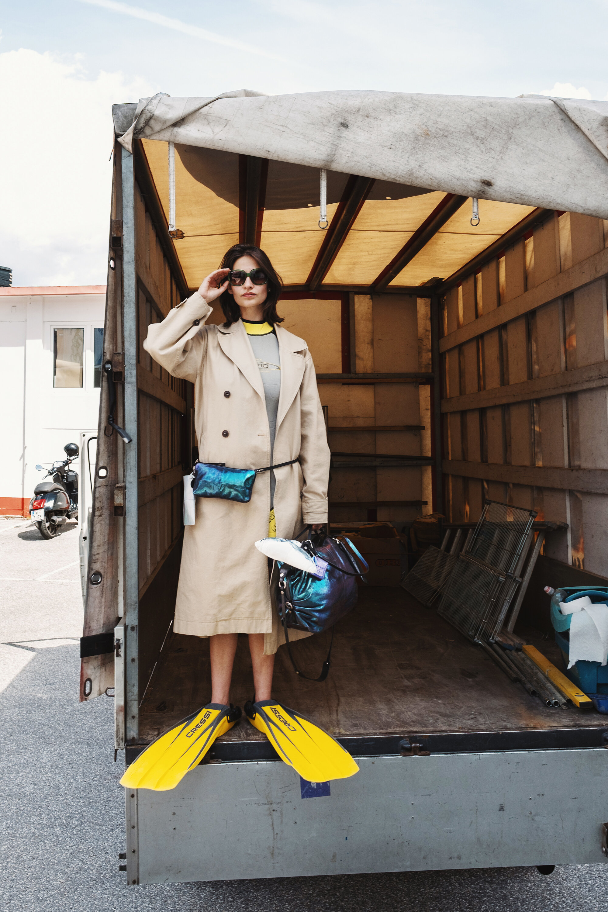 A woman in a tan trench coat, sunglasses, and yellow swim fins stands in the back of an open truck, holding a metallic INA KENT bag.