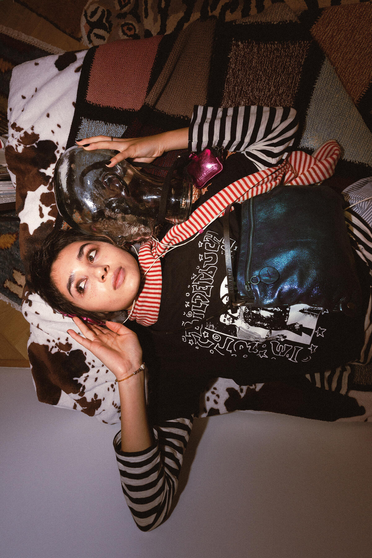 A person with striped sleeves lies on a patterned pillow, holding a glass skull and a metallic INA KENT bag, wearing a black graphic shirt and a red-and-white scarf.