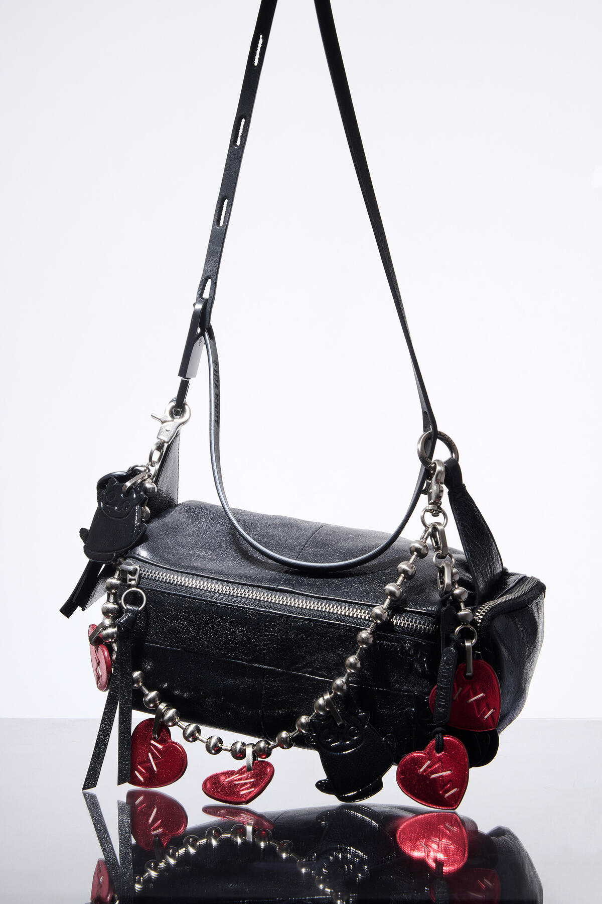 Black leather INA KENT handbag with a long adjustable strap, decorated with a silver chain and red heart-shaped charms, displayed on a reflective surface against a plain background.