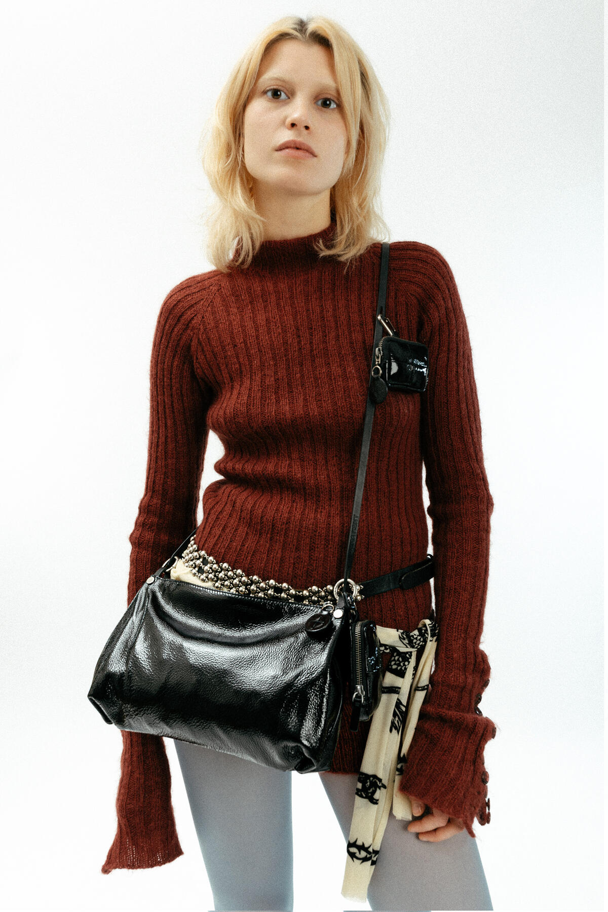 Eine Frau mit blondem Haar trägt einen kastanienbraunen Rippenpullover, eine helle Hose und eine große schwarze INA KENT-Umhängetasche mit Accessoires vor einem schlichten weißen Hintergrund.