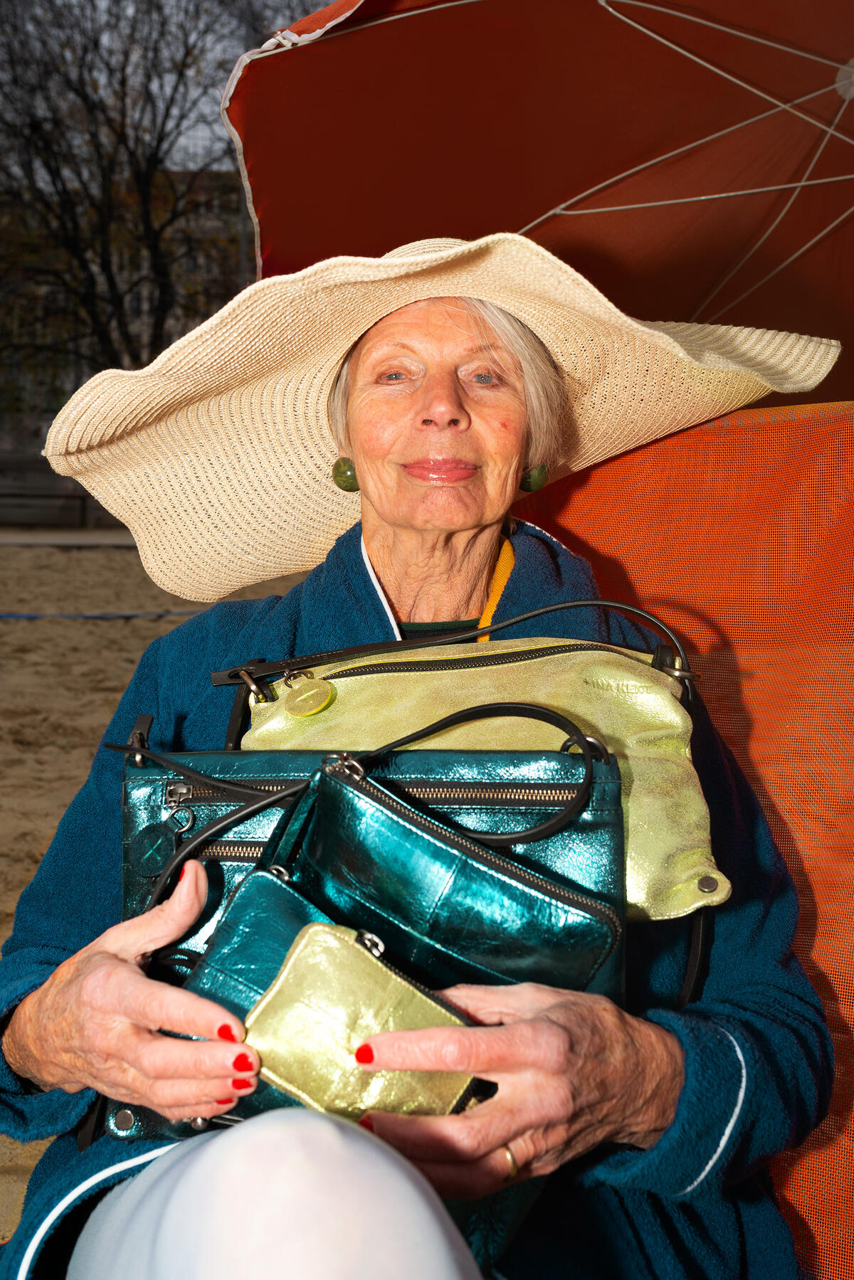 An elderly woman in a wide-brimmed hat sits outdoors, holding several shiny metallic INA KENT purses, with an orange umbrella and sandy ground in the background.