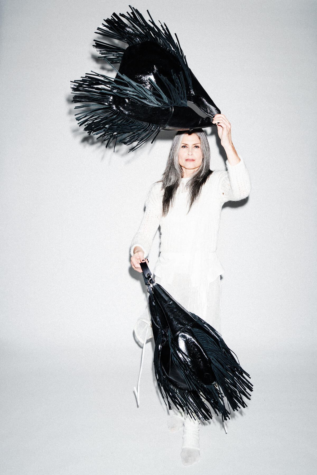 A woman in a white outfit holds and lifts two large, black, fringed INA KENT objects against a plain white background.