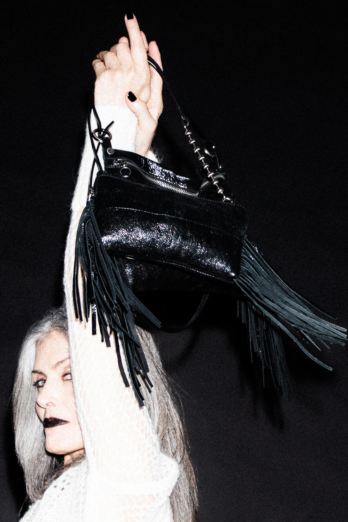 A person with long gray hair raises an INA KENT black fringed handbag against a dark background, wearing a white textured top and black lipstick.