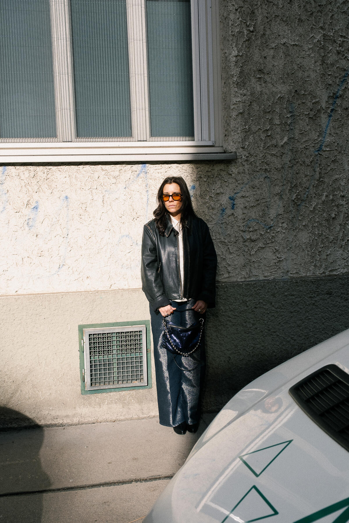 A person wearing sunglasses, a black leather jacket, and wide-leg jeans stands against a textured wall near a parked car in daylight, carrying an INA KENT bag.