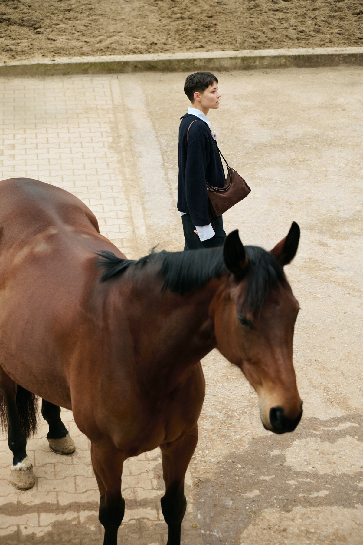 A person standing on a paved area next to a brown horse in an outdoor setting, carrying an INA KENT bag.