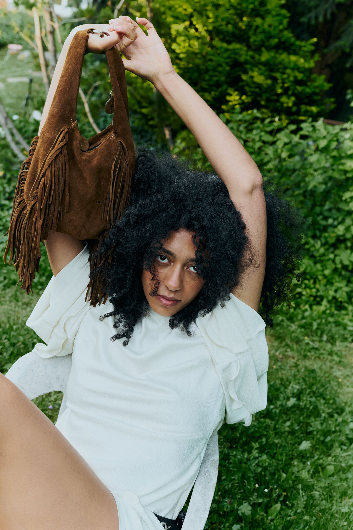 A person with curly hair sits outdoors on a white chair, holding up a brown fringed INA KENT handbag, surrounded by greenery.