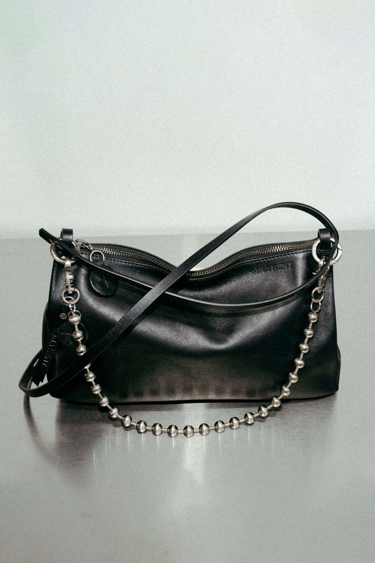 An INA KENT black leather handbag with a zipper closure, a thin shoulder strap, and a decorative silver beaded chain, placed on a reflective surface against a plain background.